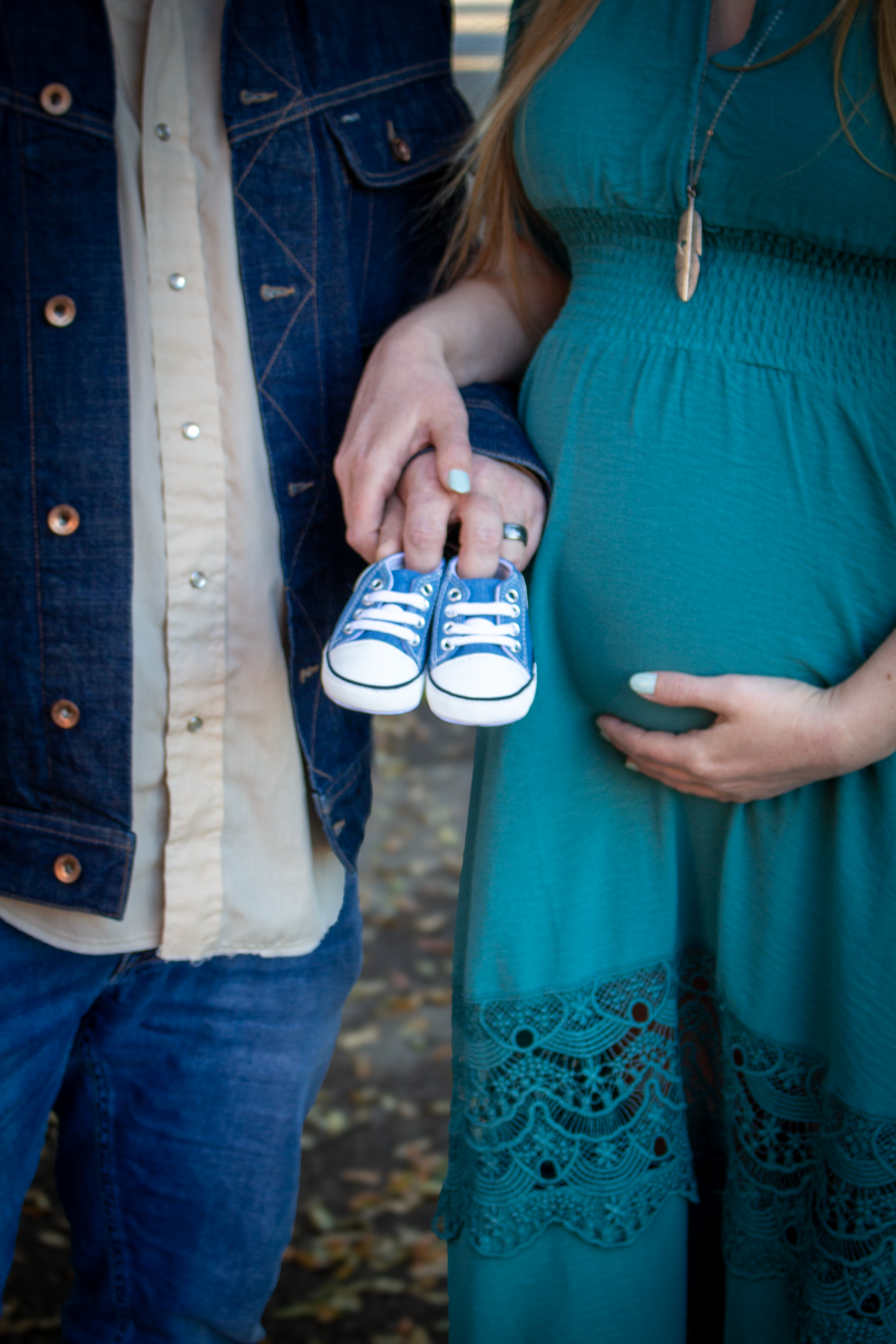 A pregnant woman in a teal dress holds tiny blue baby shoes while standing next to someone in denim.