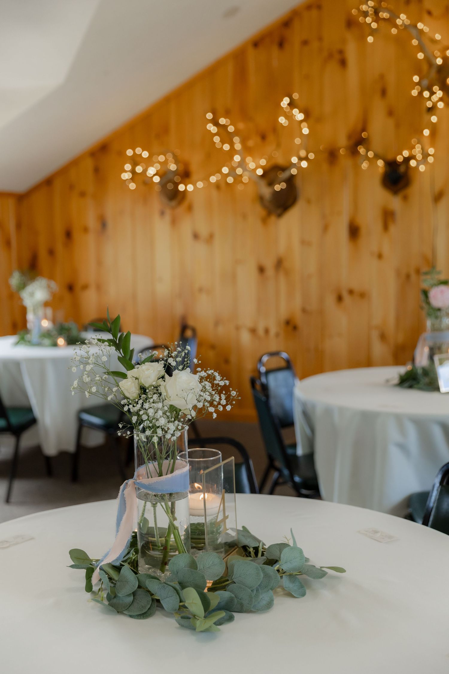 Simple wedding reception centerpiece with white flowers and eucalyptus on round table in rustic wood-paneled venue with twinkle lights.