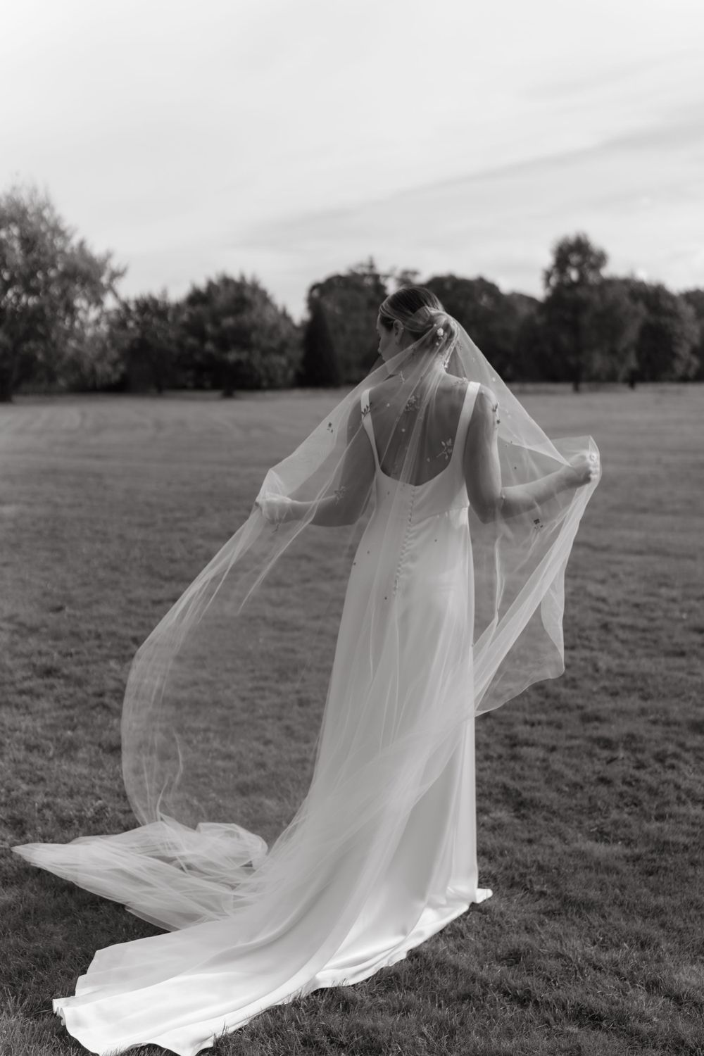 A back view of a bride in a flowing white wedding dress with a delicate veil billowing in the wind on a grassy field.