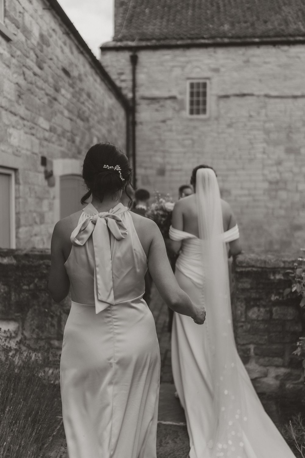 A black and white wedding photo showing elegant dresses with flowing gowns and veils against a stone building wall.