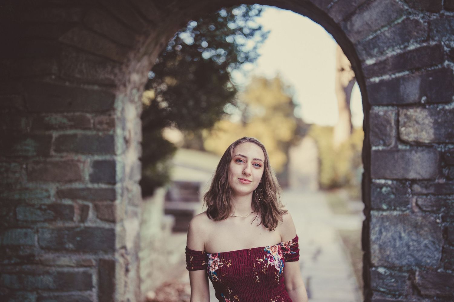 A woman in a burgundy off-shoulder dress stands under a stone archway during golden hour.