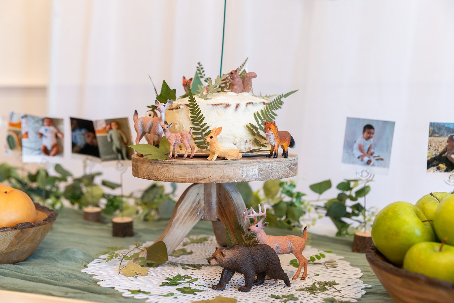 A decorated white cake on a wooden stand with a toy bear and greenery displayed near fresh green apples.