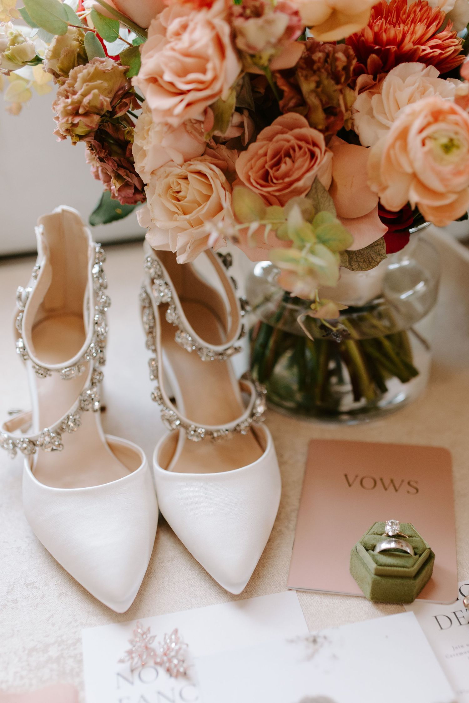 Elegant white bridal shoes with crystal details beside pink roses and vows book.