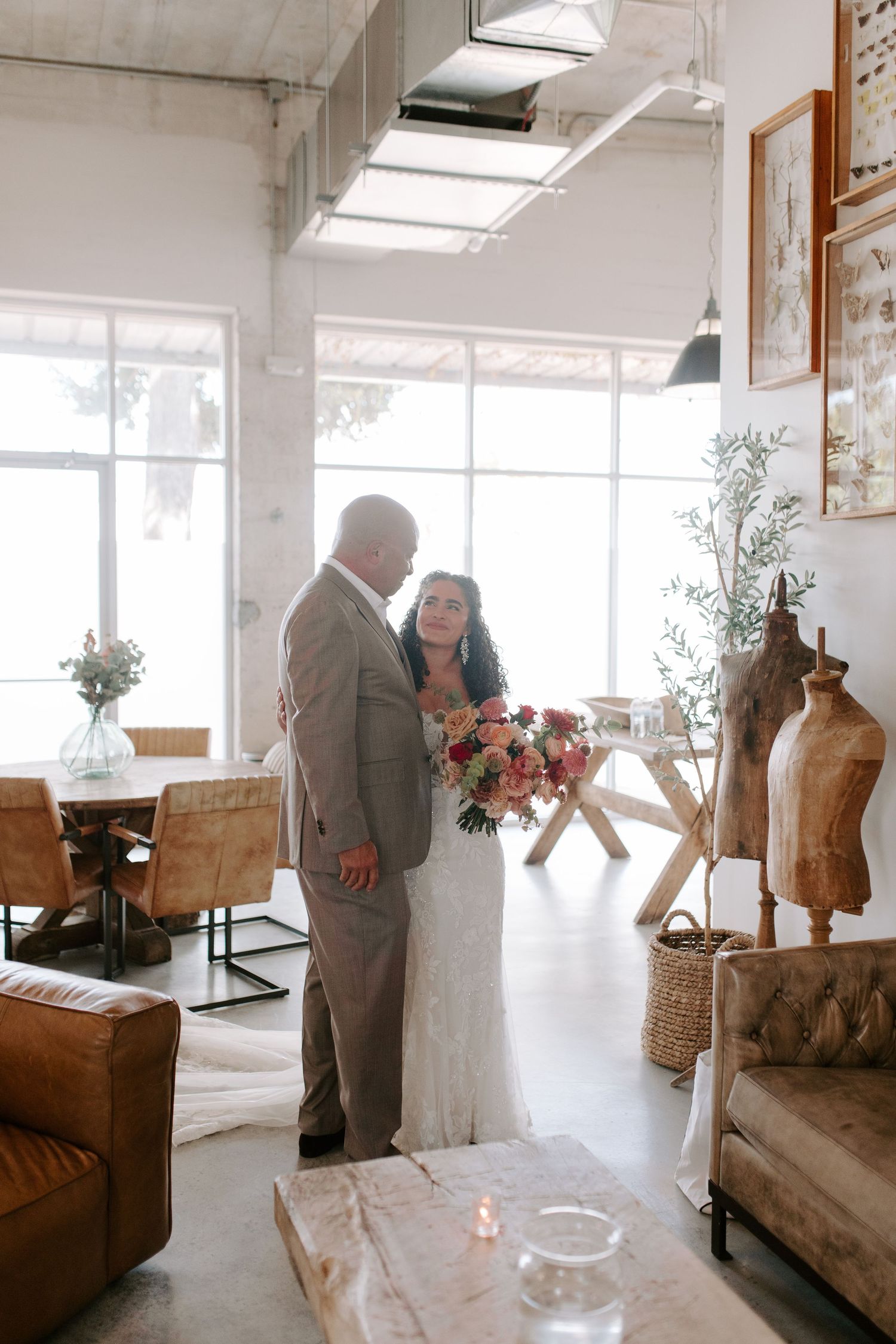 A couple in wedding attire share an intimate moment in a bright industrial-style venue with vintage decor.