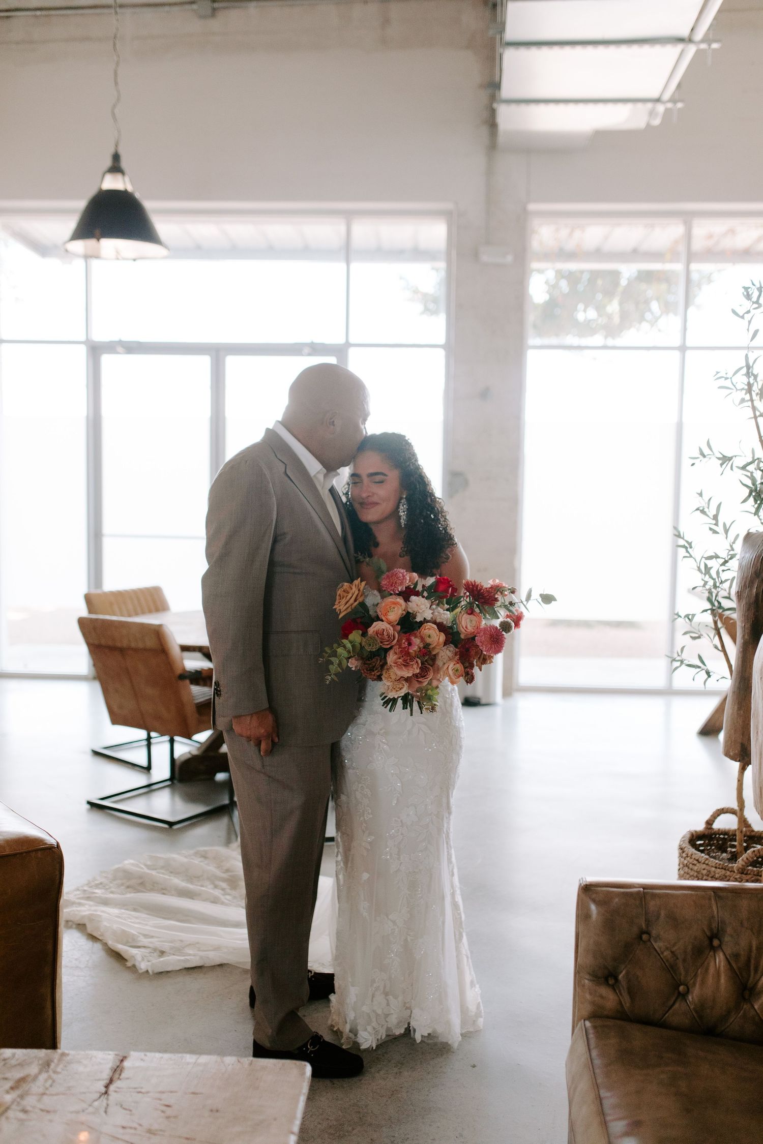 A wedding couple stands together in a sunlit room with modern decor and warm tones.