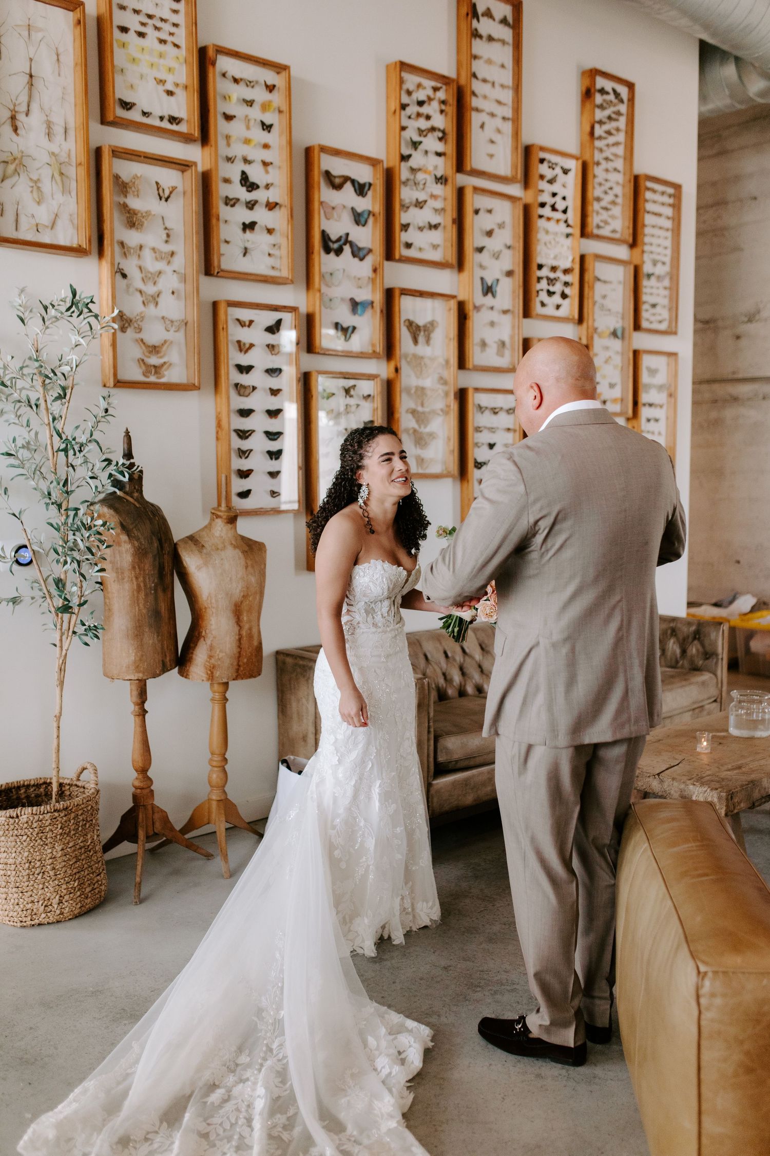 A couple in wedding attire stands by a wall of framed butterfly displays in an industrial space.
