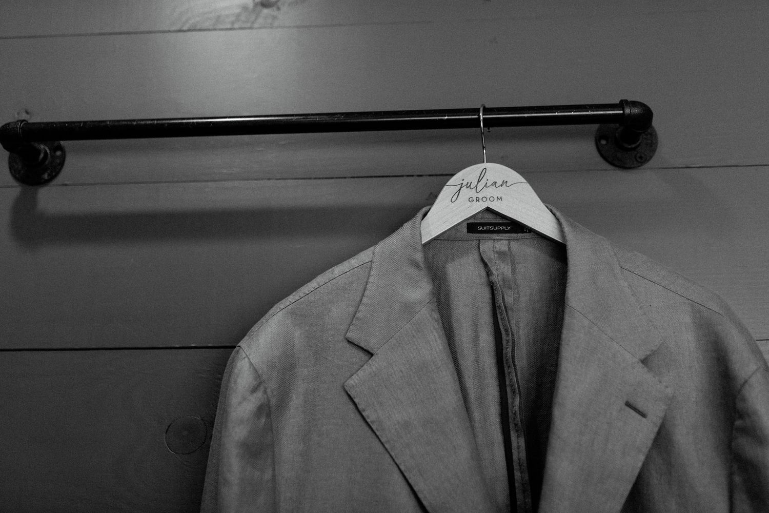 A gray suit jacket hangs on a black pipe hanger against a wall.