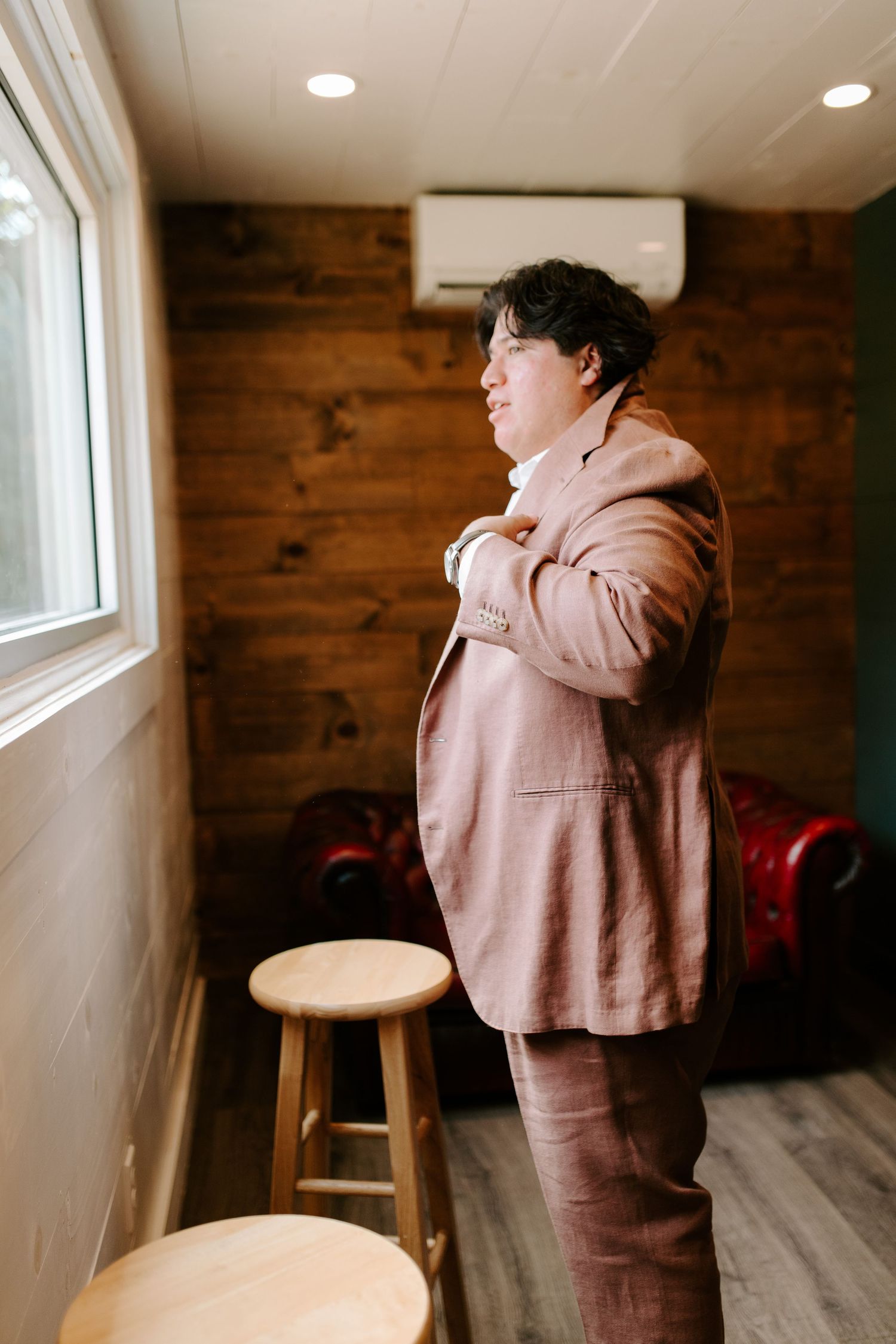 Person in a pink suit adjusts their collar while standing near a window in a wood-paneled room.