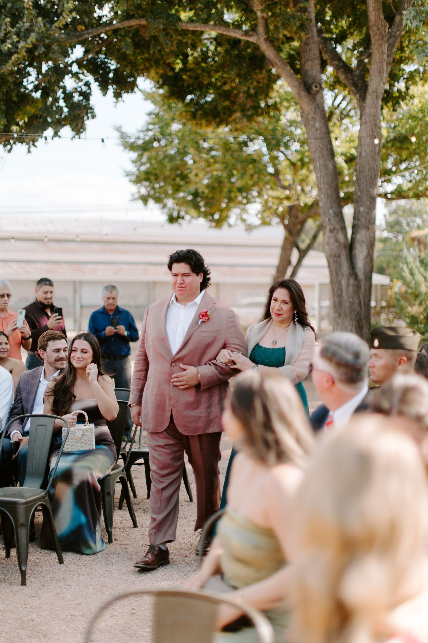 A wedding ceremony processional featuring a couple walking down an aisle lined with guests under shady trees.