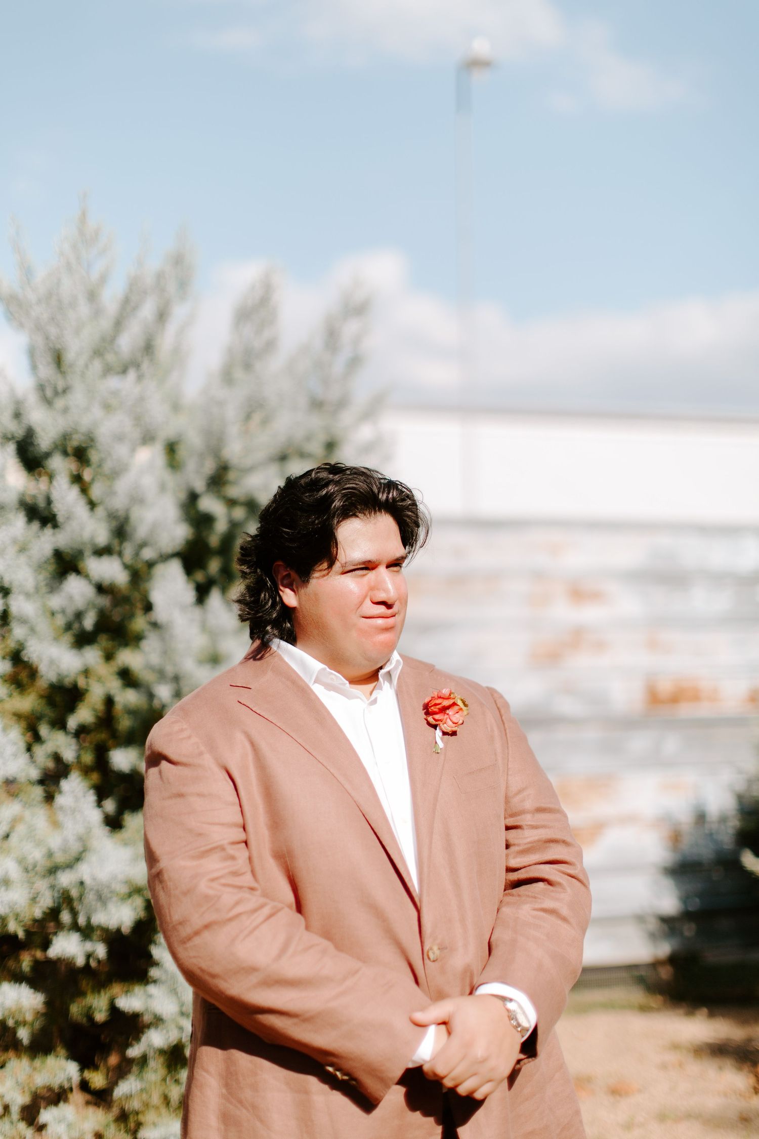 A groom in a dusty pink suit jacket stands outdoors on a sunny day.