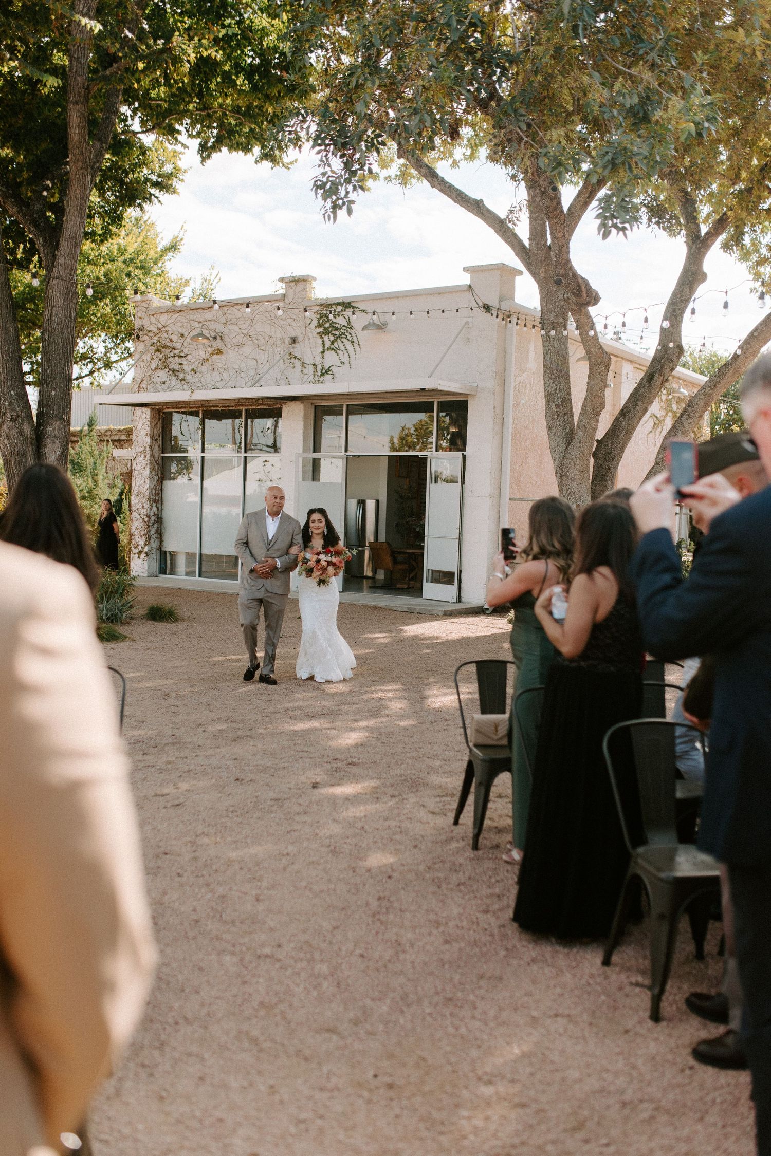 An outdoor wedding ceremony with guests seated on black chairs in front of a modern white building.