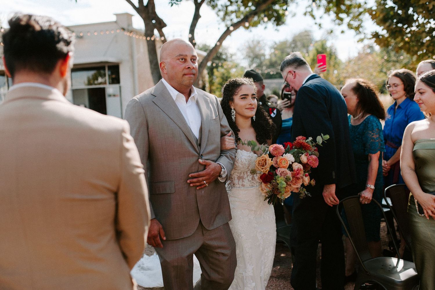A wedding processional moment outdoors with guests gathered under trees.
