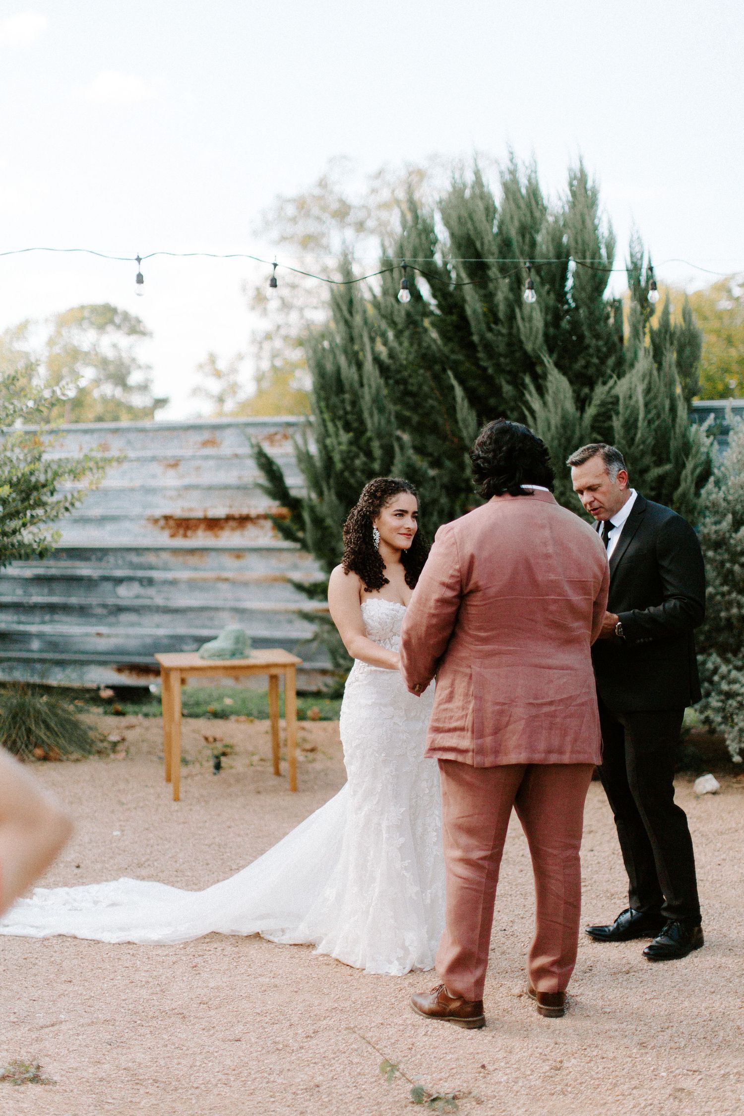 A wedding ceremony taking place in an outdoor garden setting with string lights overhead.