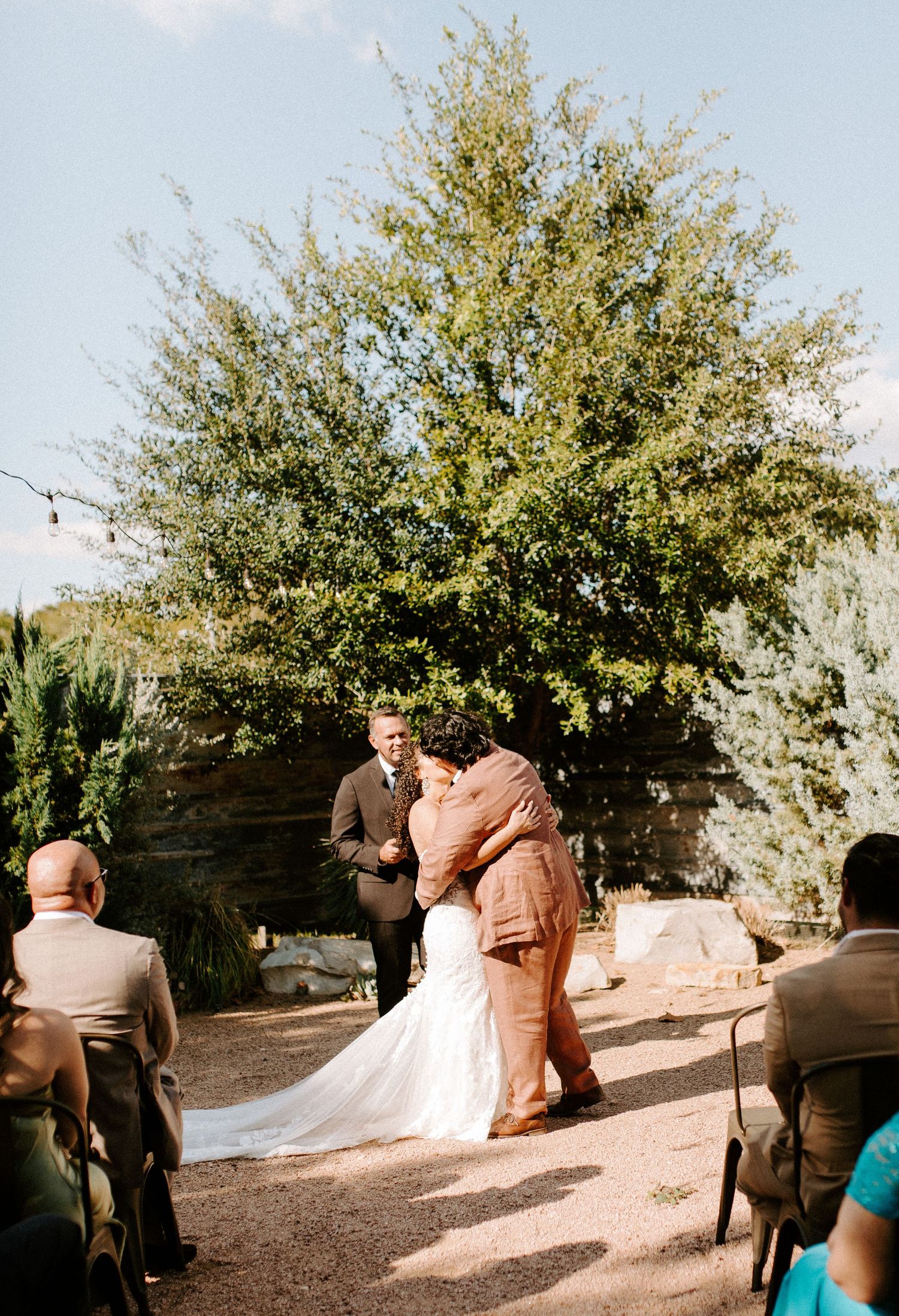 A wedding ceremony takes place under a large tree with guests seated on folding chairs.