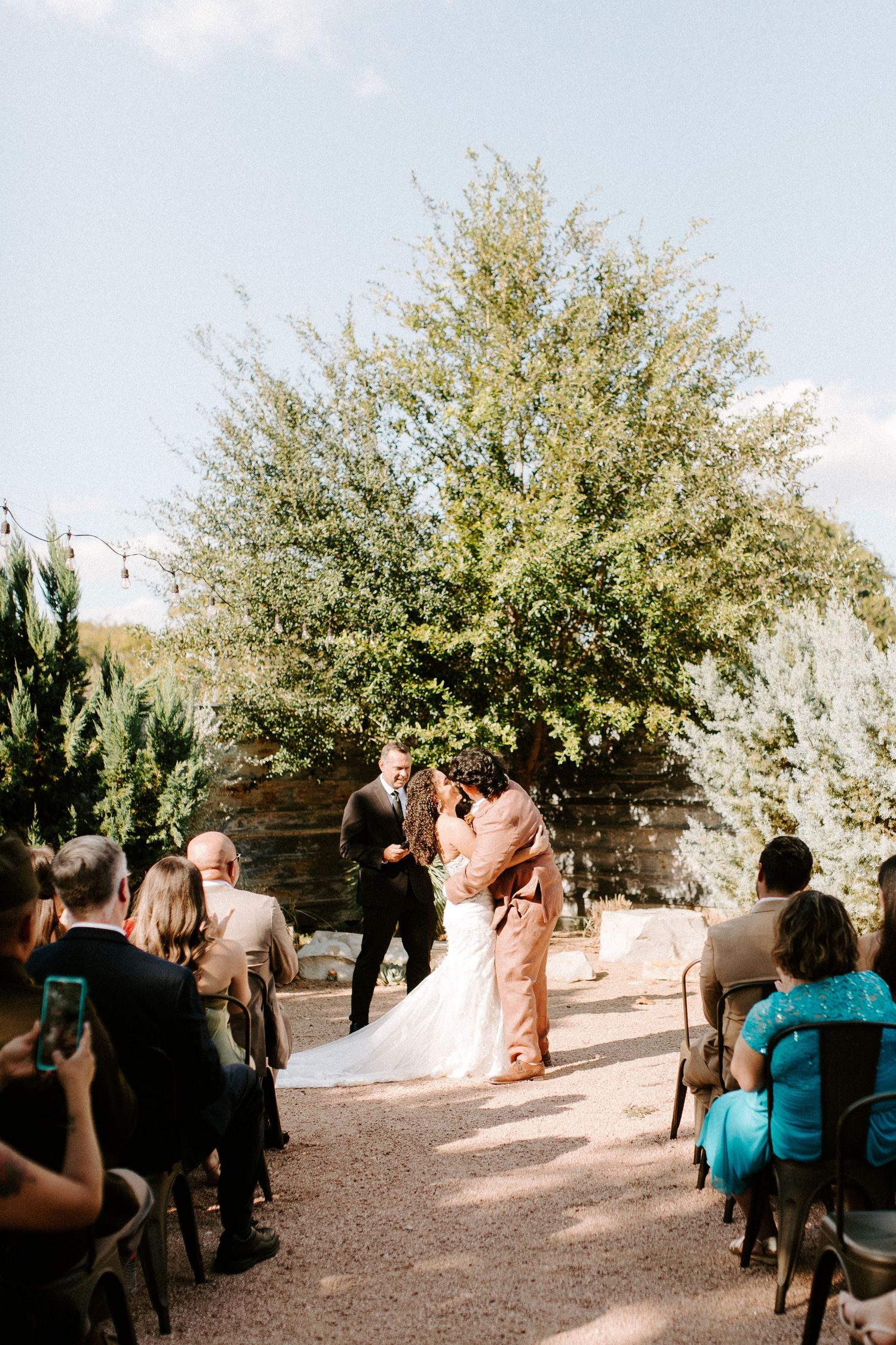 An outdoor wedding ceremony beneath a sprawling tree with guests watching the couple.