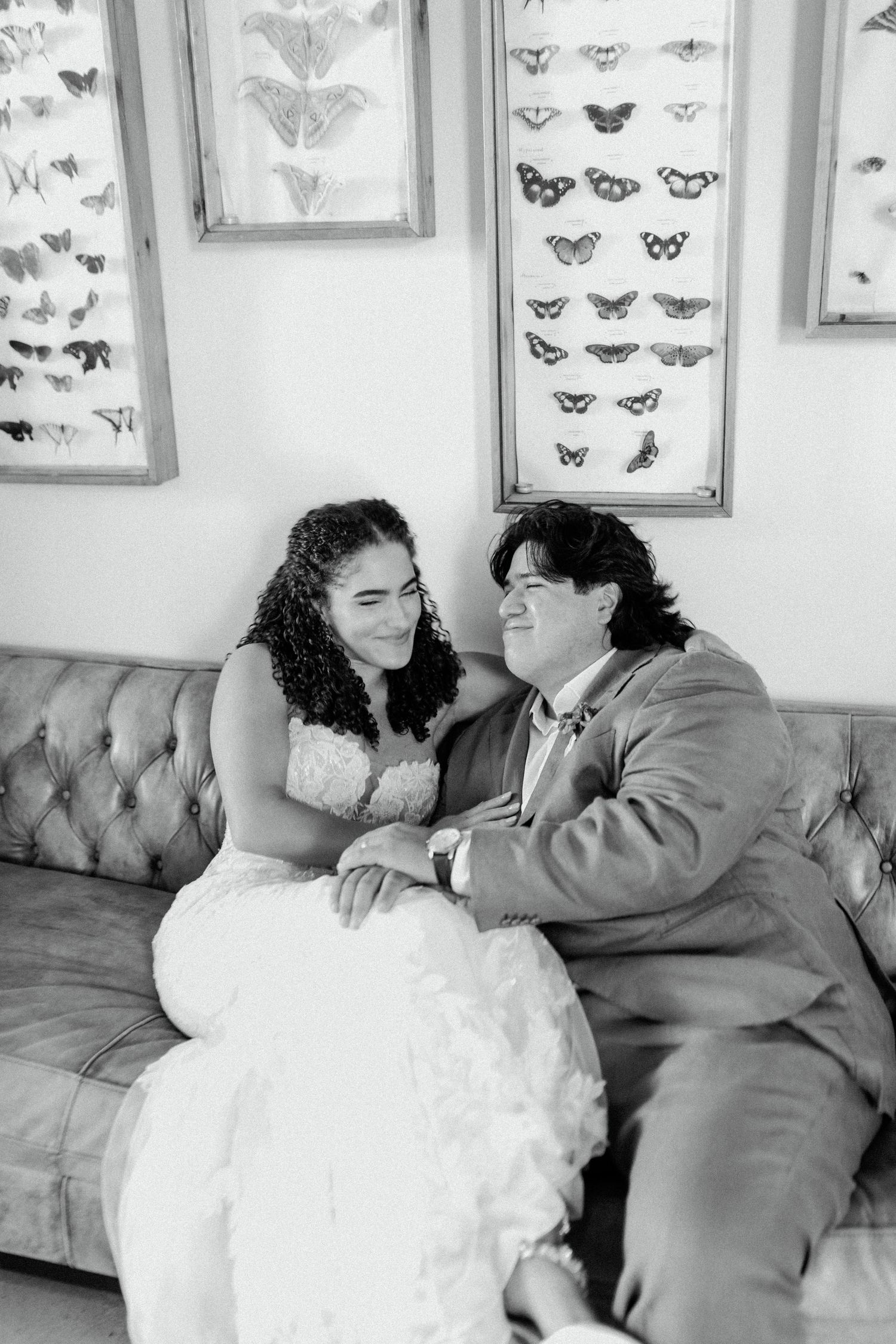 A couple shares a tender moment on a couch with framed butterfly displays on the wall.