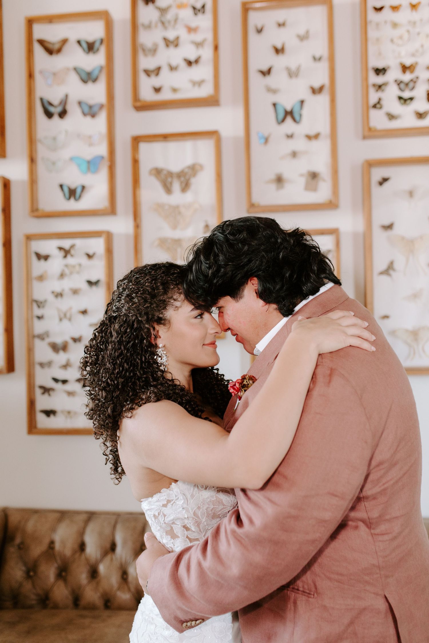 Wedding couple embraces in front of butterfly artwork display on beige wall for romantic indoor portrait.