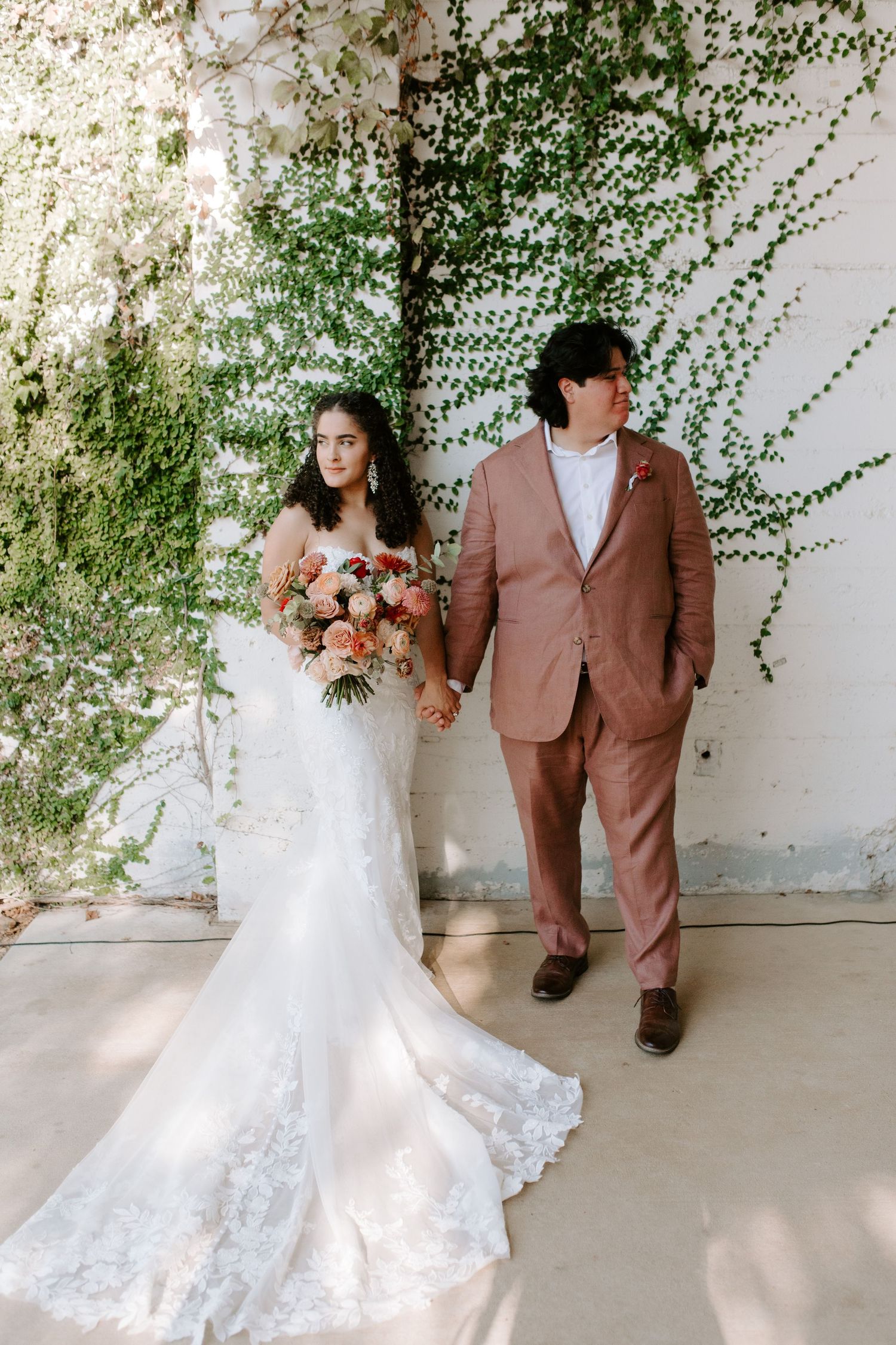 Bride in lace gown and groom in dusty rose suit pose against vine-covered wall.