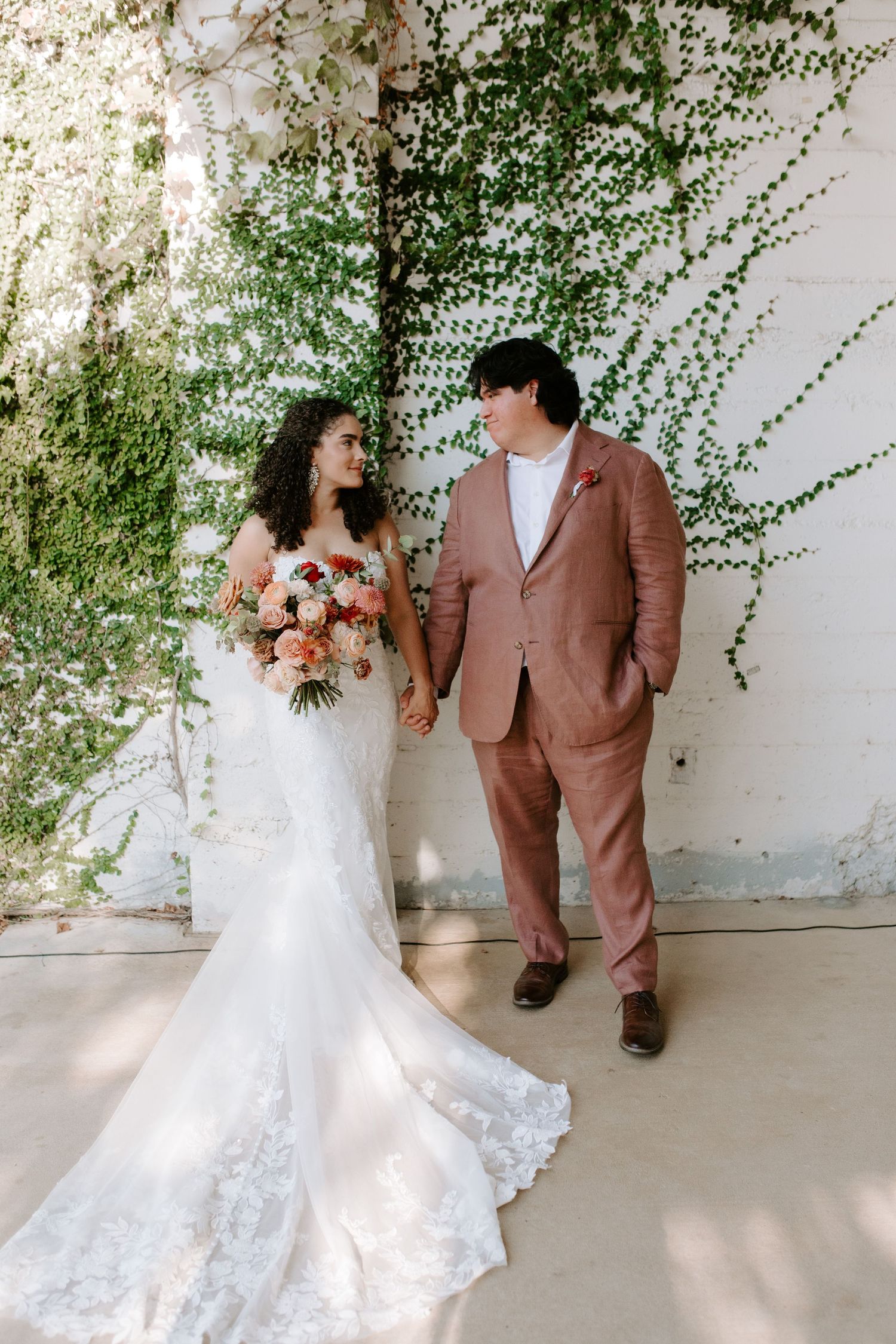 Couple in wedding attire standing by ivy wall, bride wearing detailed lace gown.