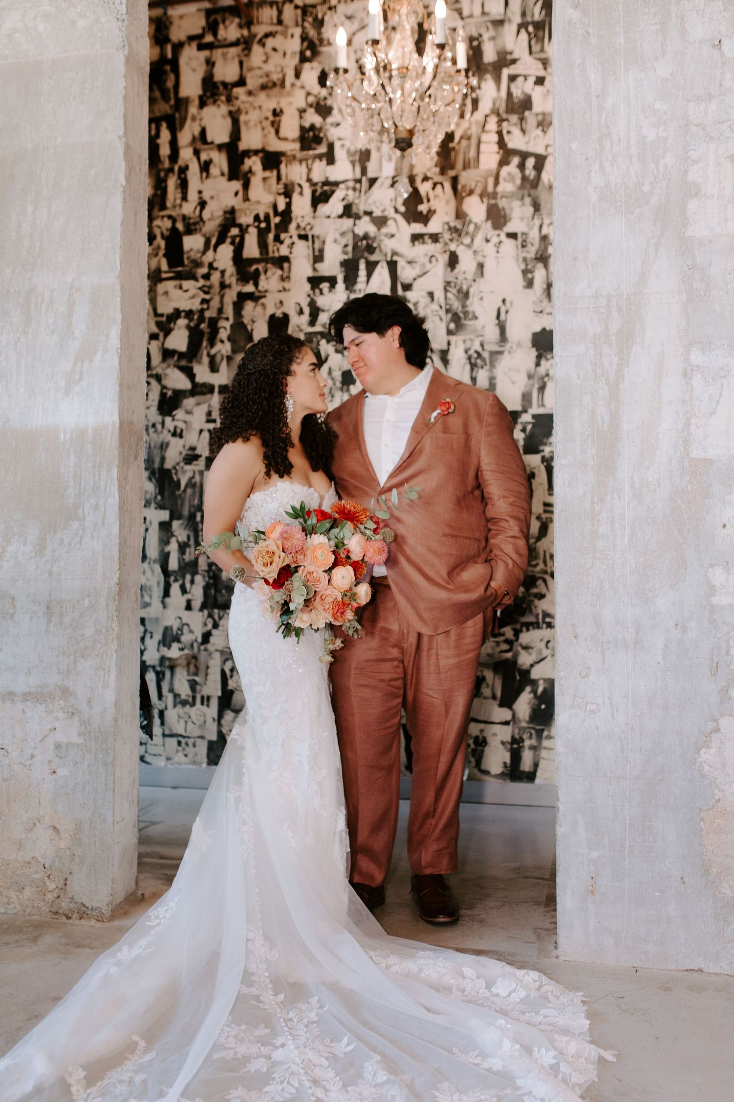 Elegant couple poses against textured wall, bride in lace gown and groom in rose-colored suit.