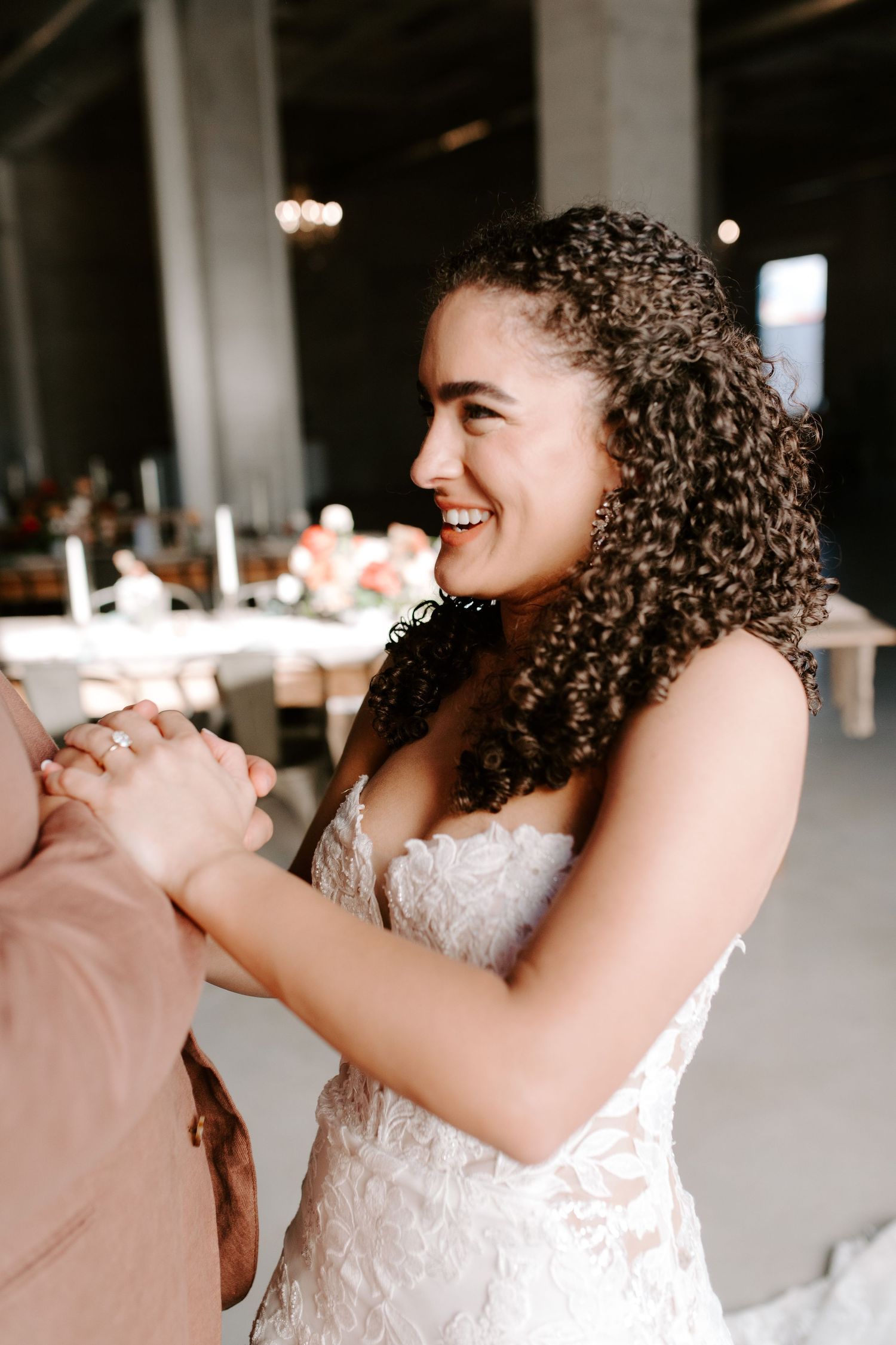Bride in lace gown smiles radiantly while dancing with partner in modern venue.