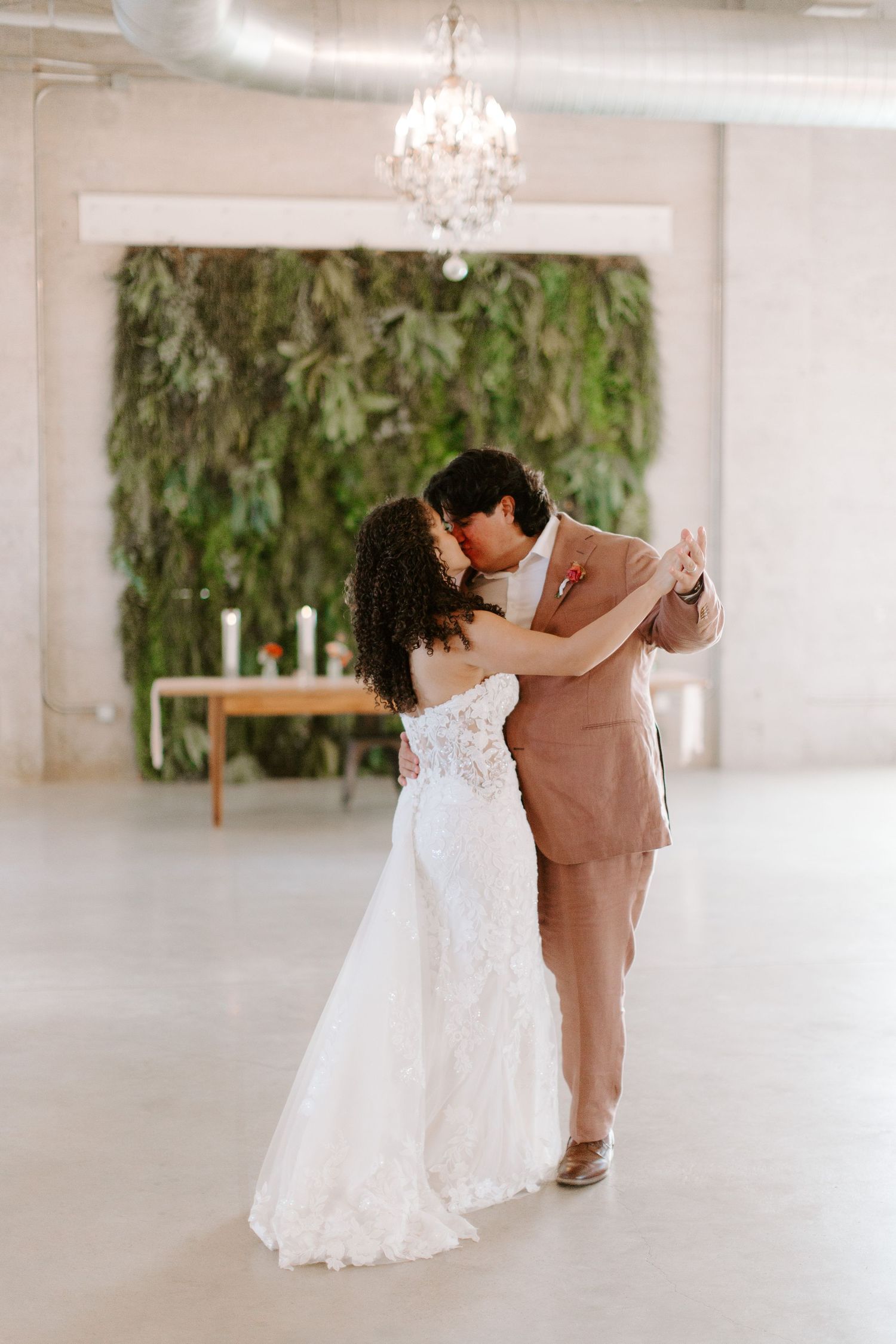 Wedding couple dances beneath chandelier with lush greenery wall backdrop.