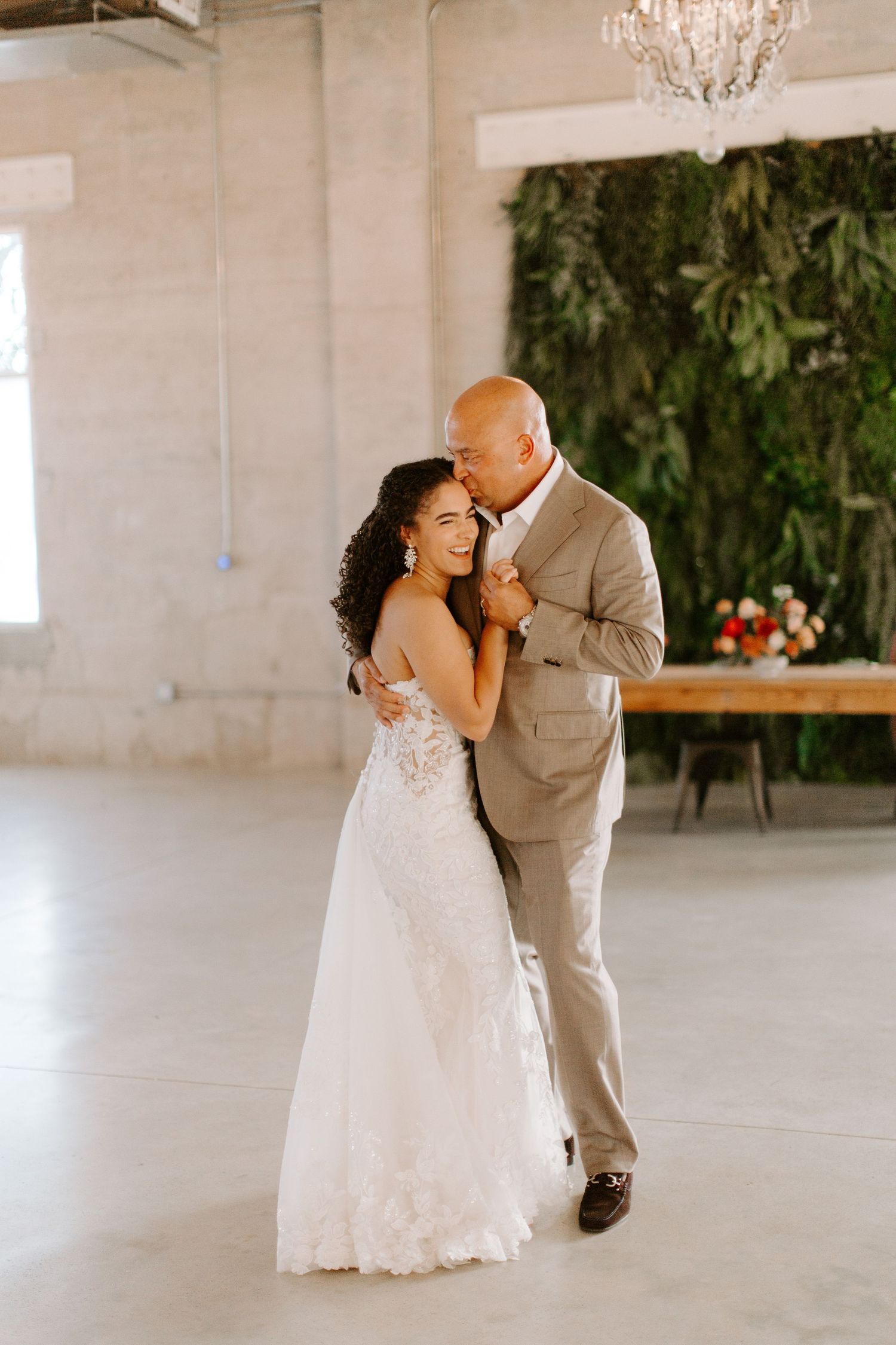 Bride in lace gown dances with partner against modern white venue with greenery wall.