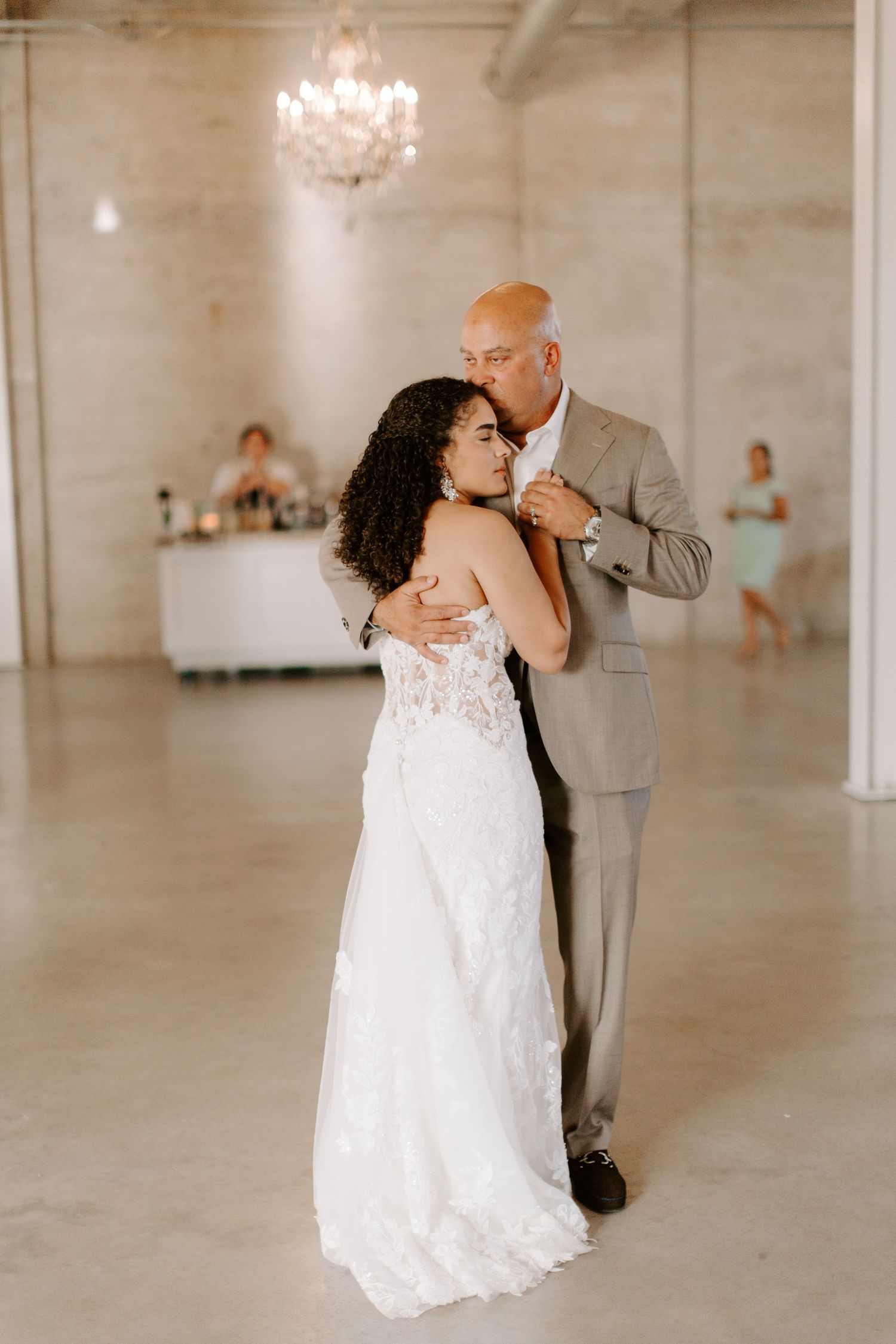 A tender moment between newlyweds dancing in a white wedding dress and gray suit under crystal chandeliers.