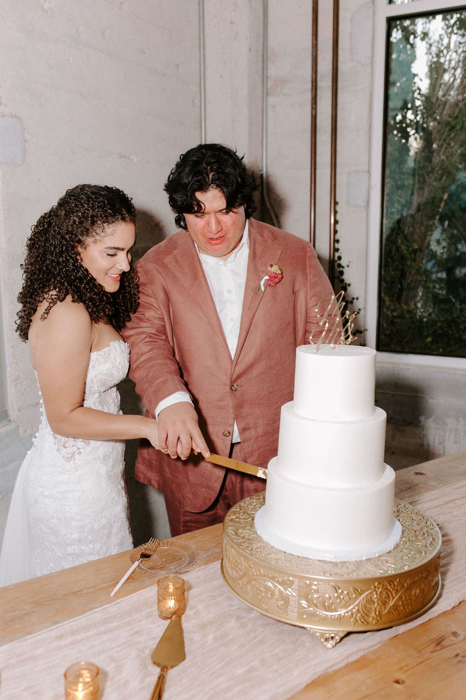 Newlyweds cut their three-tiered white wedding cake together on a decorative gold stand.