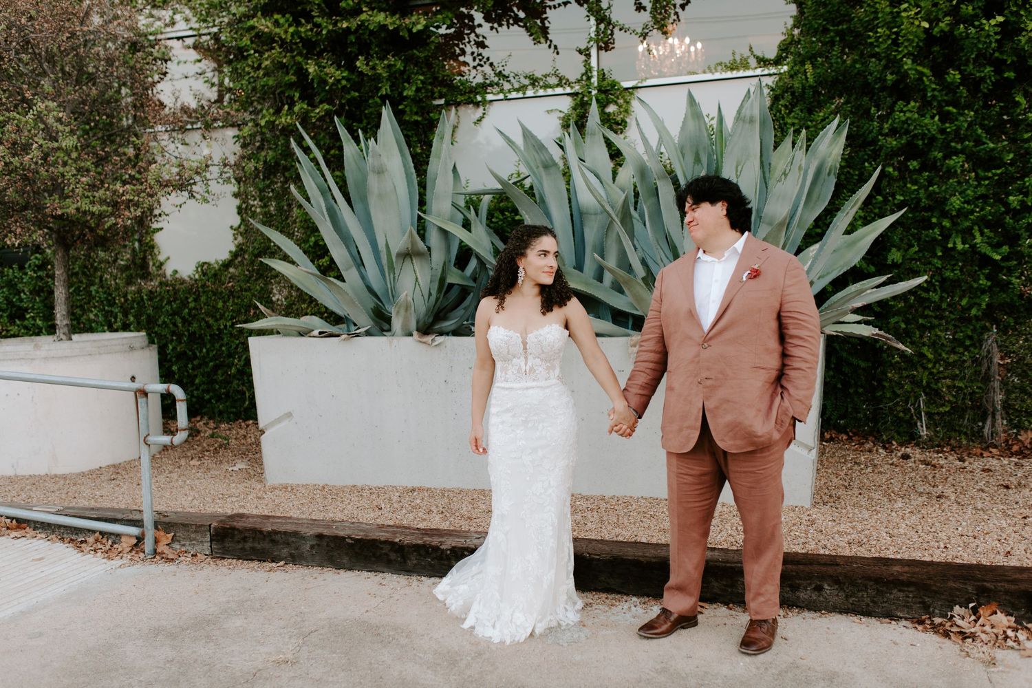 Newlyweds hold hands while posing in front of desert landscaping.