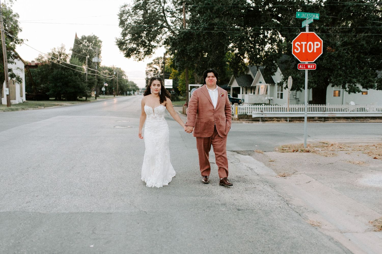A couple stands on a residential street in front of a stop sign, with the person wearing a brown suit and white wedding dress.
