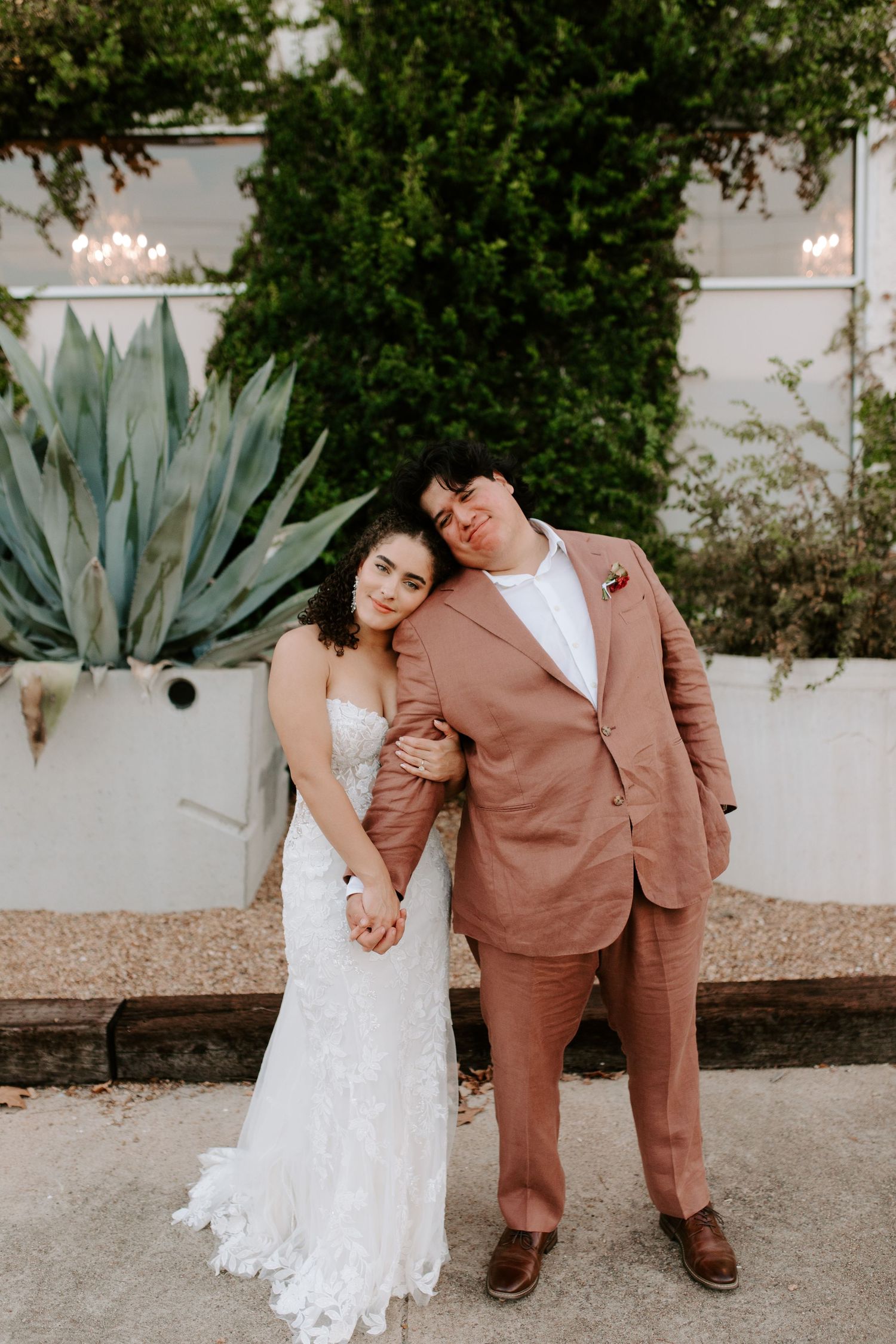 Wedding couple poses by white wall and agave plant with groom in salmon colored suit.
