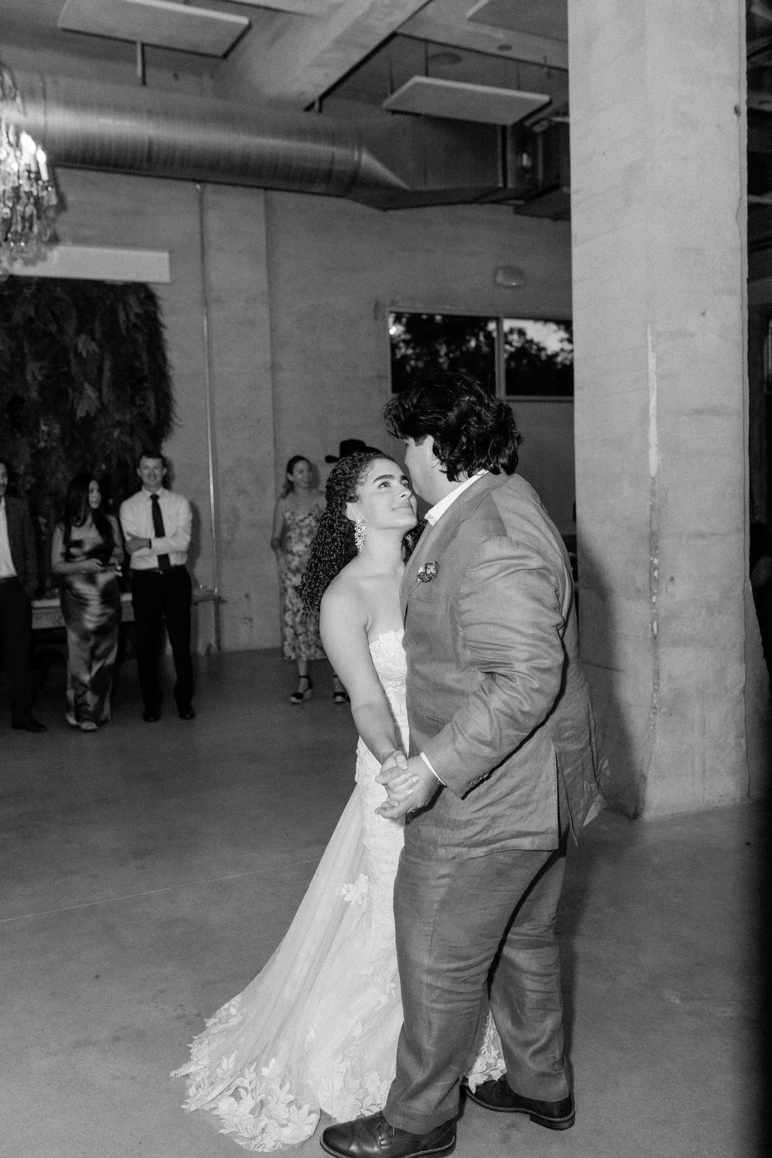 Black and white photo of couple dancing at indoor reception venue with industrial decor.
