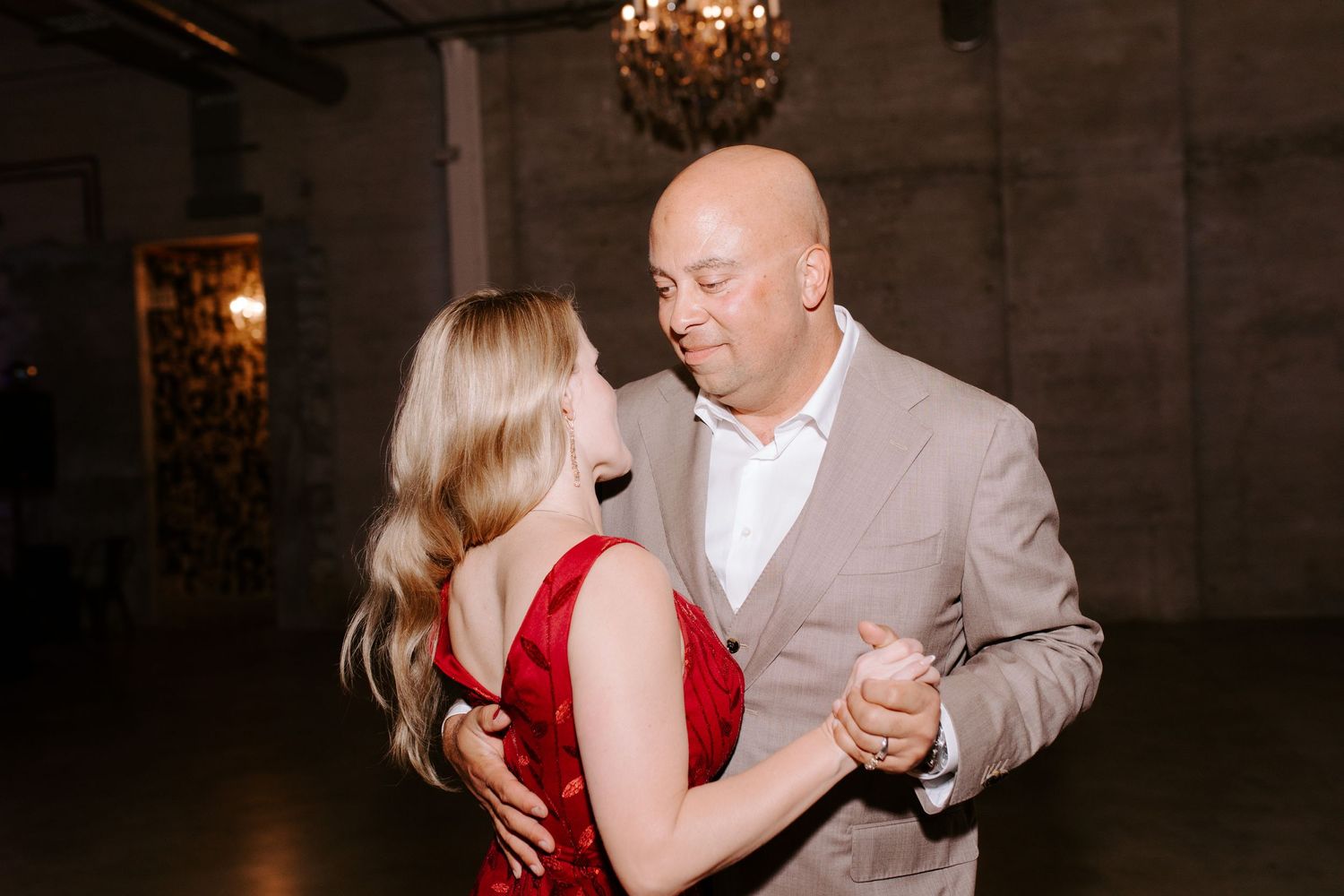 A couple shares an elegant dance together in a dimly lit venue with a chandelier overhead.