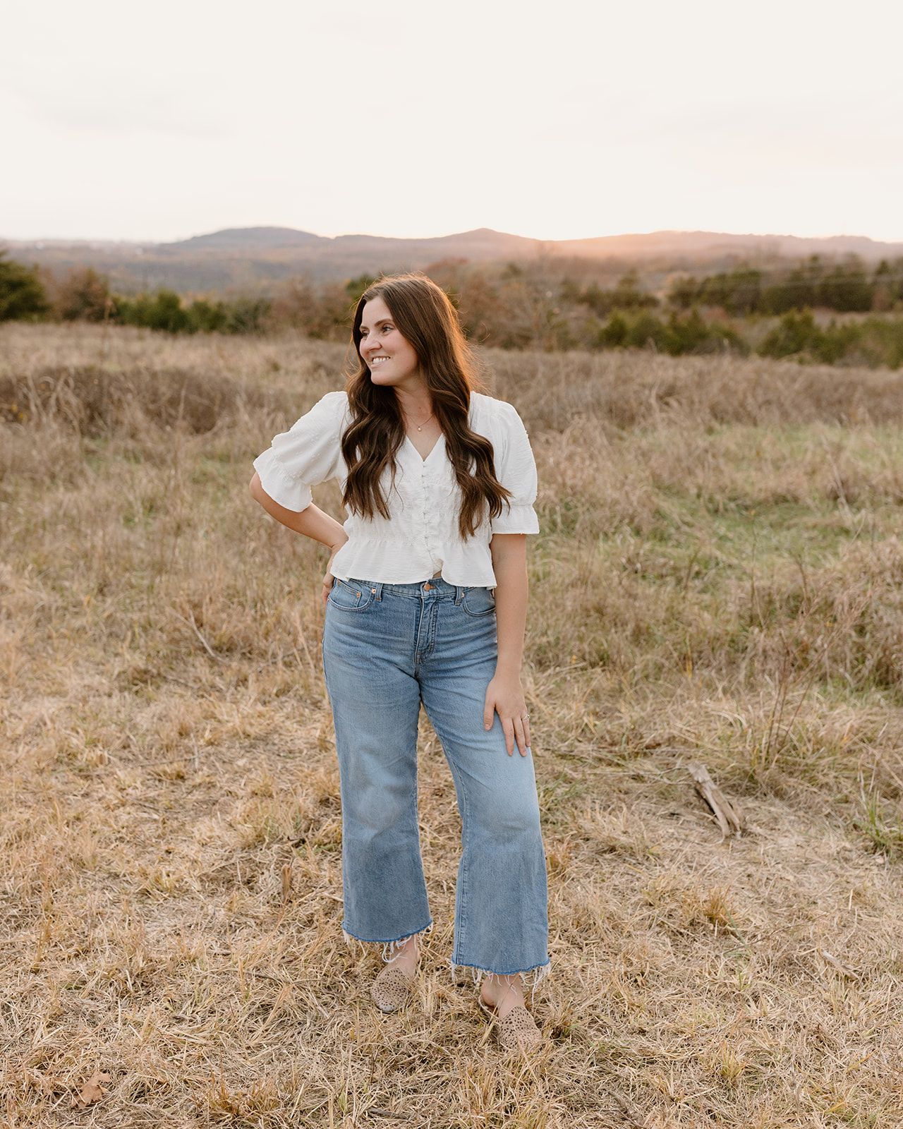 A woman in jeans and white top poses in a field with mountains at sunset.