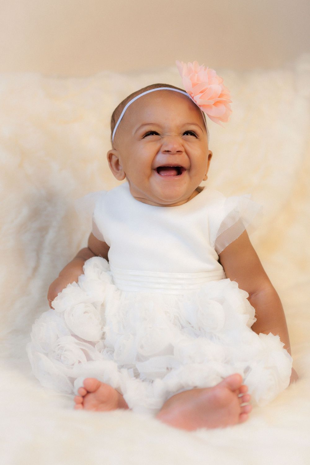 Joyful baby wearing white dress and peach flower headband sits on soft fur blanket with bright smile.