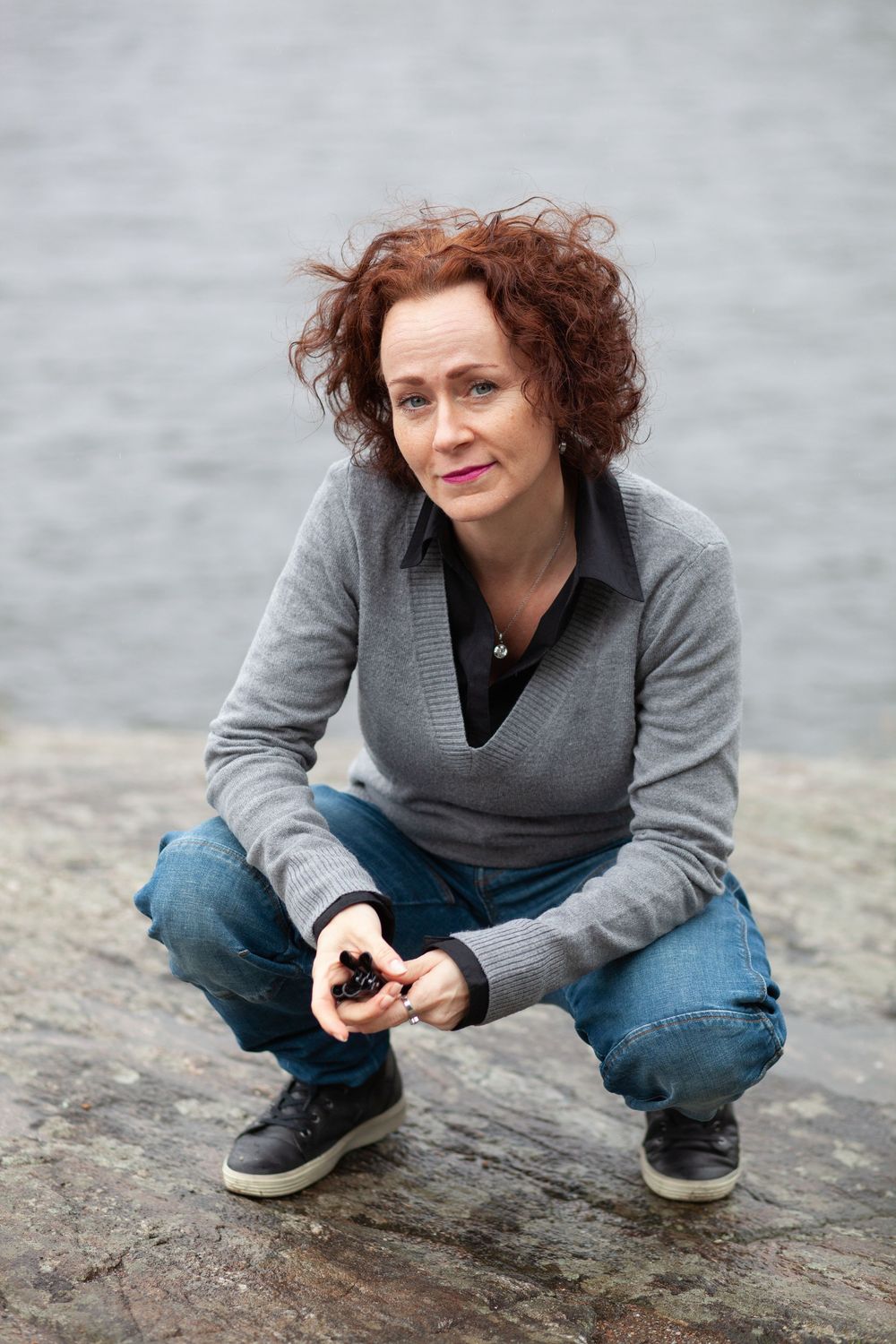 Person wearing a gray sweater and jeans crouches on a rocky surface near water.