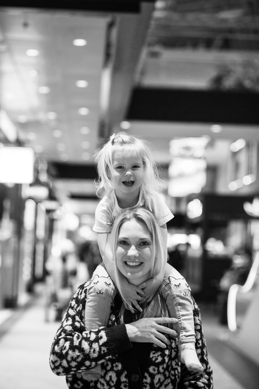 Black and white candid photo showing playful moment with people smiling in a shopping mall corridor.