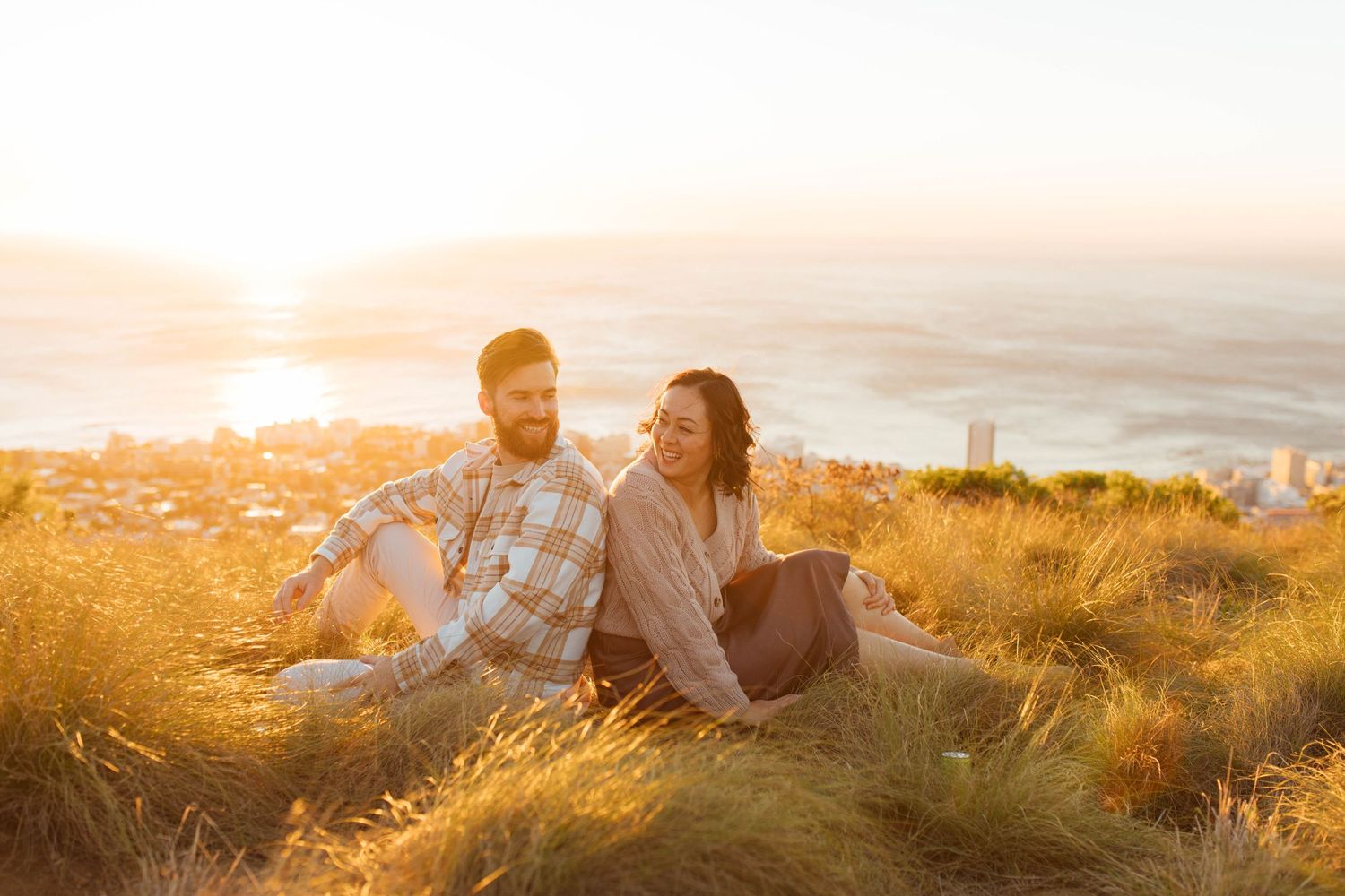 Couple sitting together on grassy hilltop at sunset overlooking coastal cityscape in golden light.