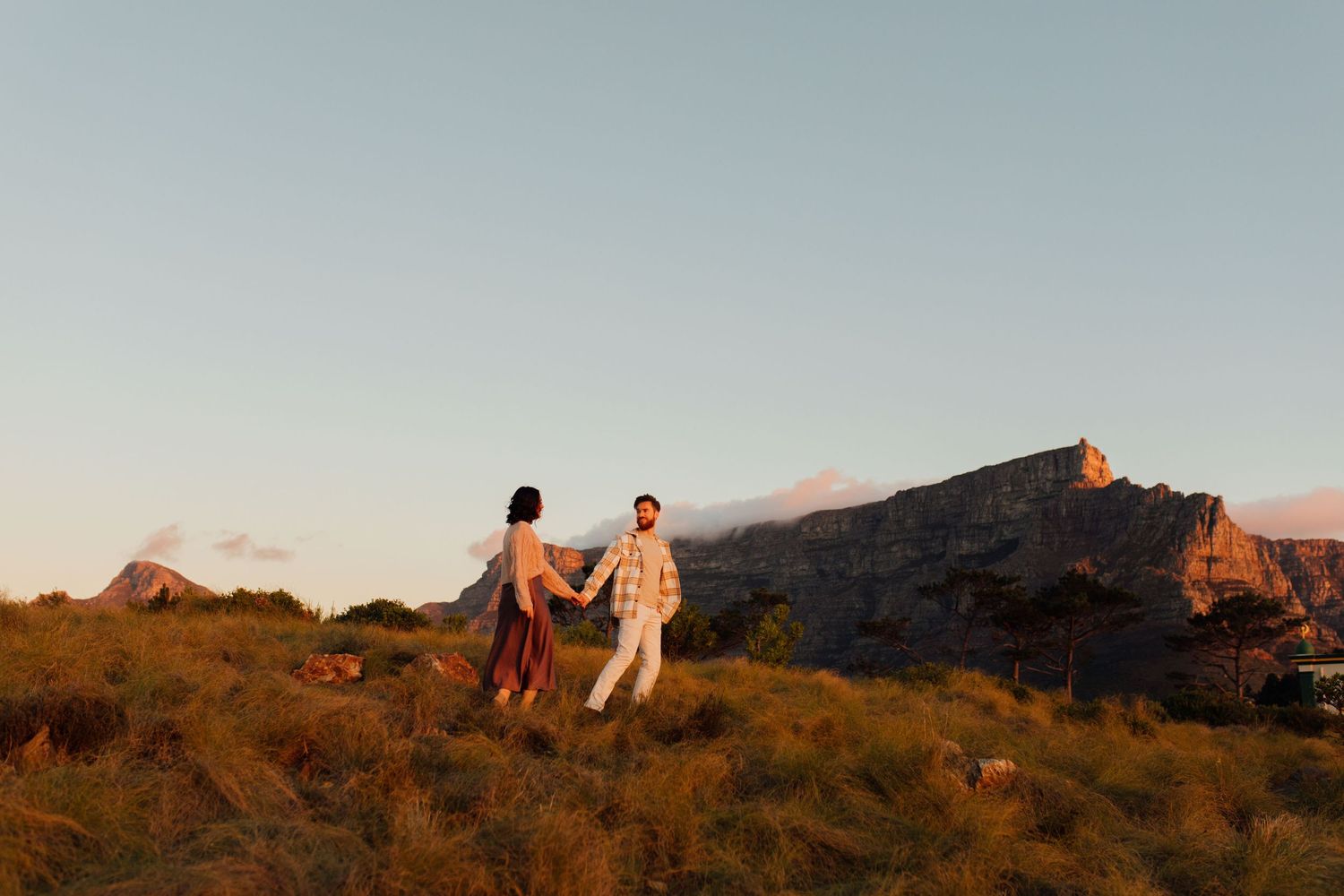 Two people walking together at sunset on a mountainside with golden light illuminating the rugged landscape and peaks.