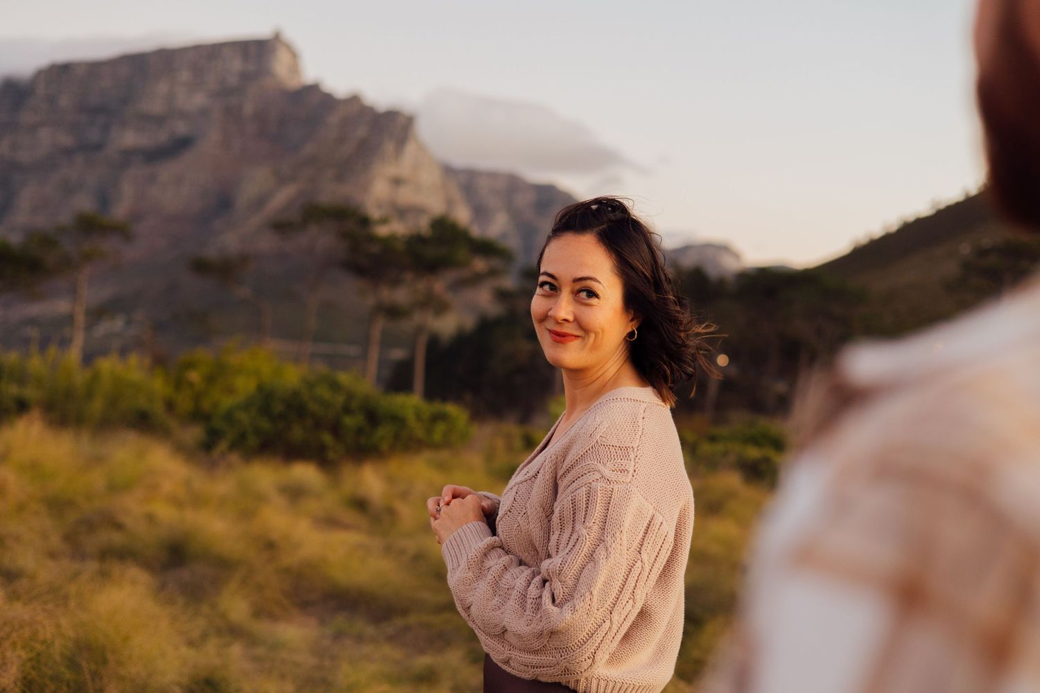 A person in a beige sweater stands before a scenic mountain landscape at sunset in warm golden light.