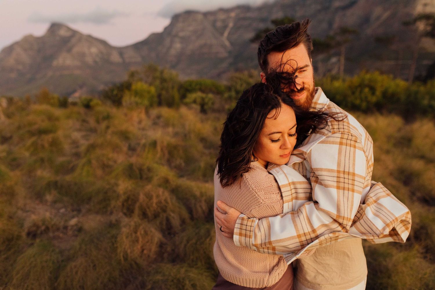 Two people embrace while wrapped in a cozy plaid blanket against a backdrop of Table mountain landscapes at sunset.
