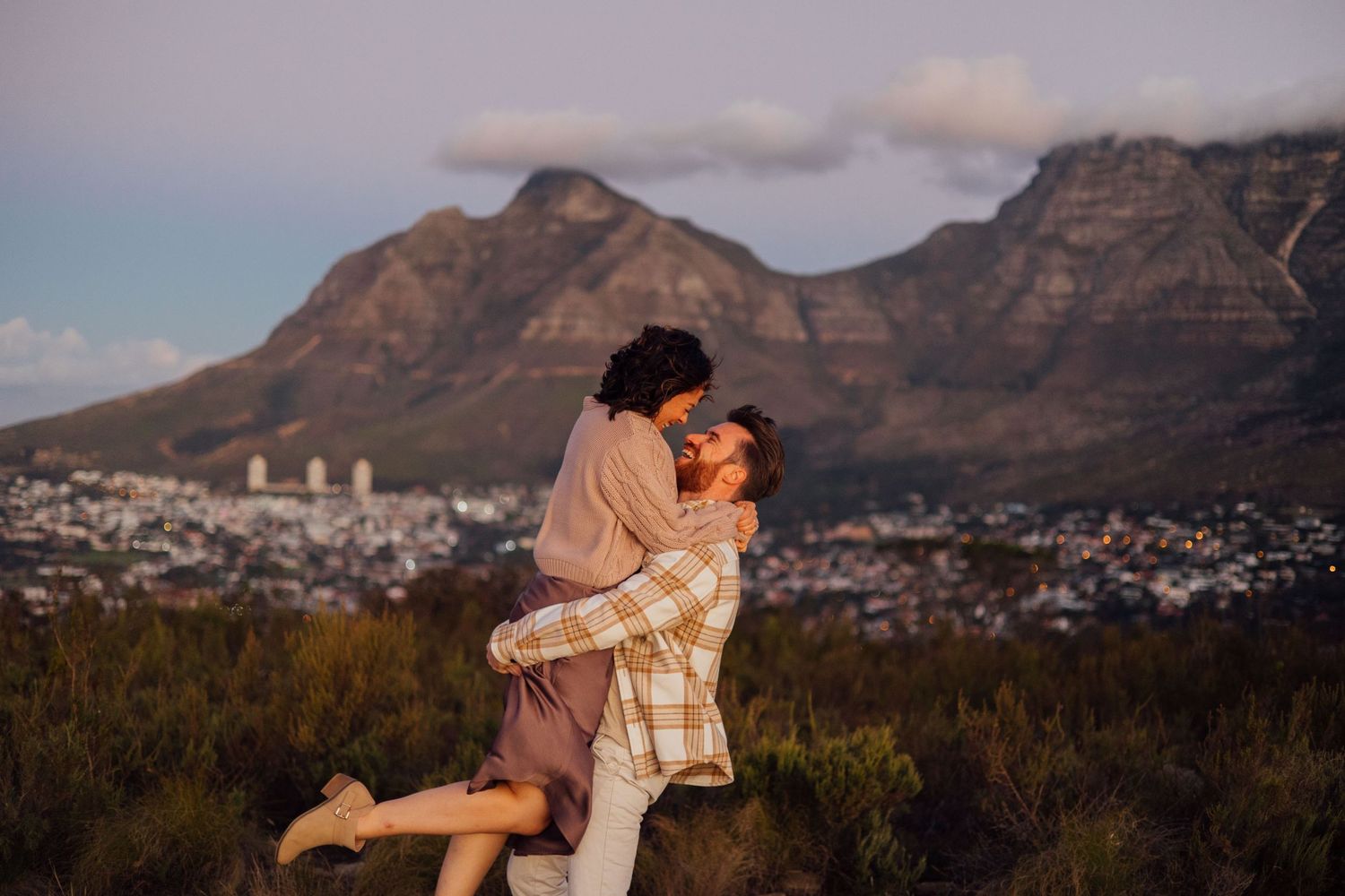 A romantic couple embraces against the backdrop of Table Mountain in Cape Town during golden hour sunset.