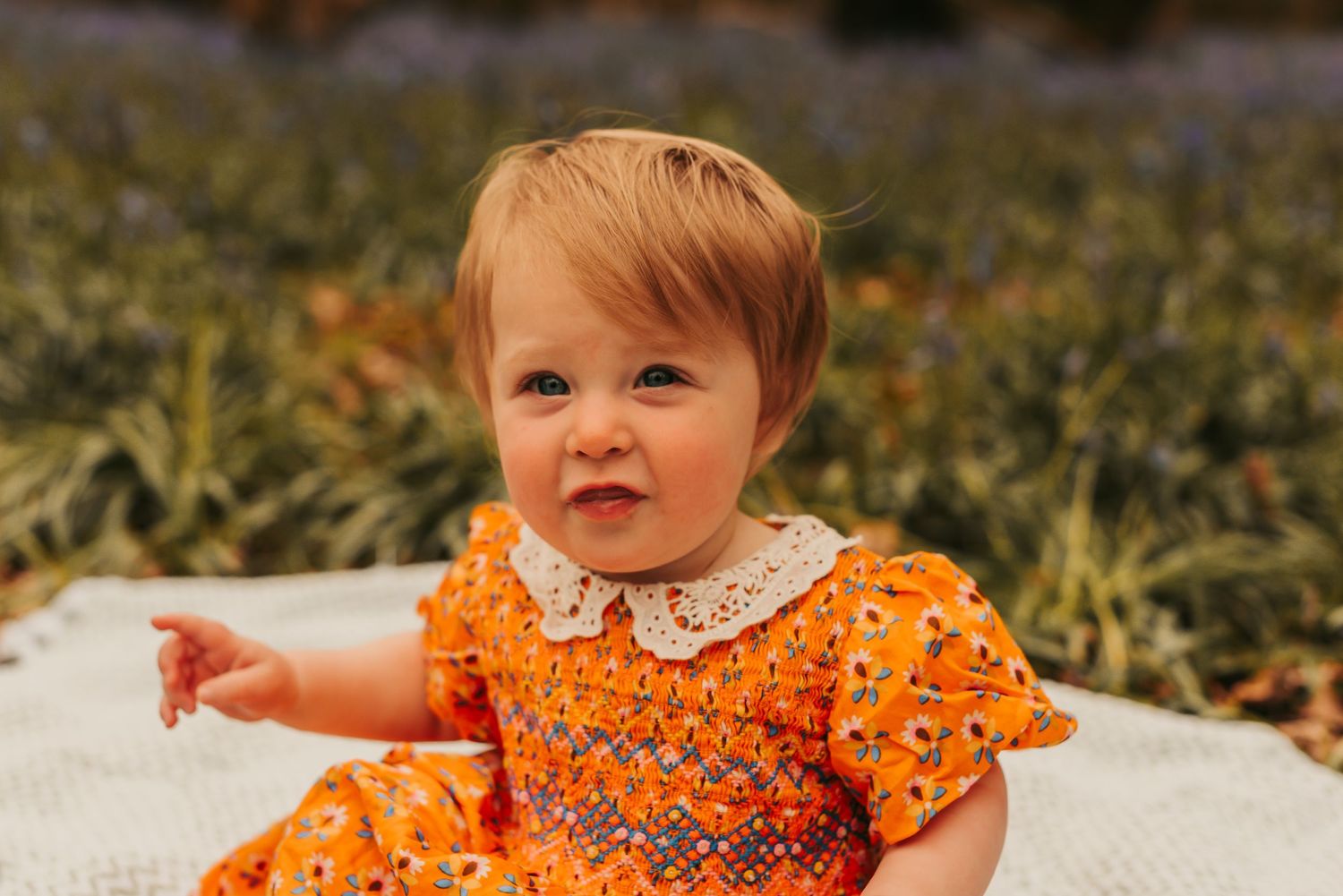 Small toddler sits on blanket outdoors wearing an orange floral dress with white lace collar during autumn.