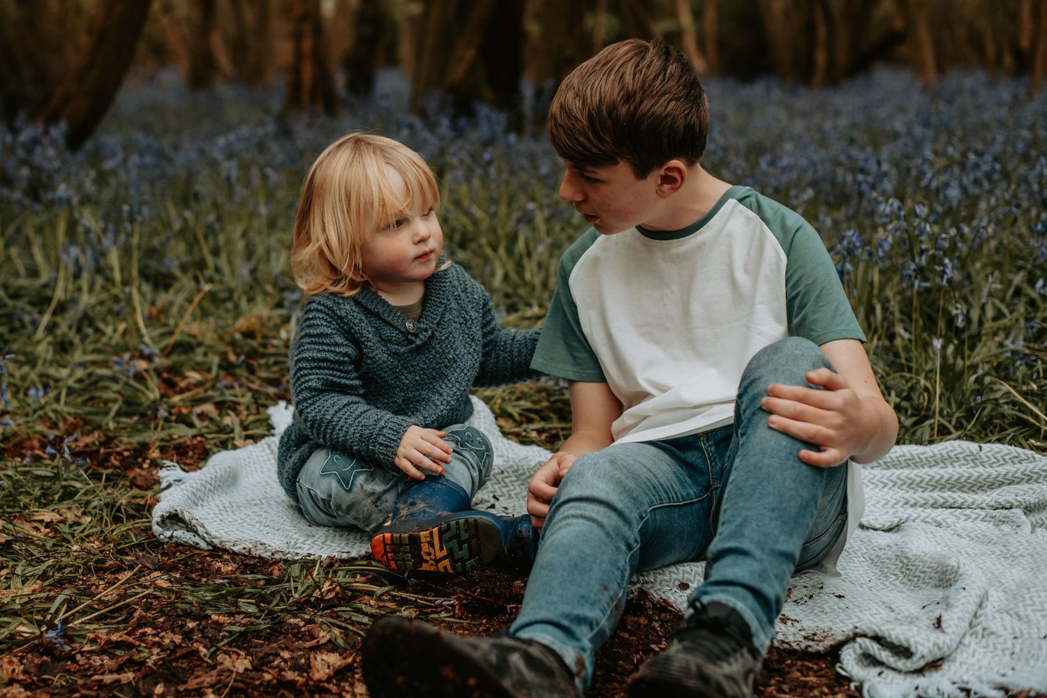 Two children sit together on a blanket in a woodland setting with blooming bluebells in spring.