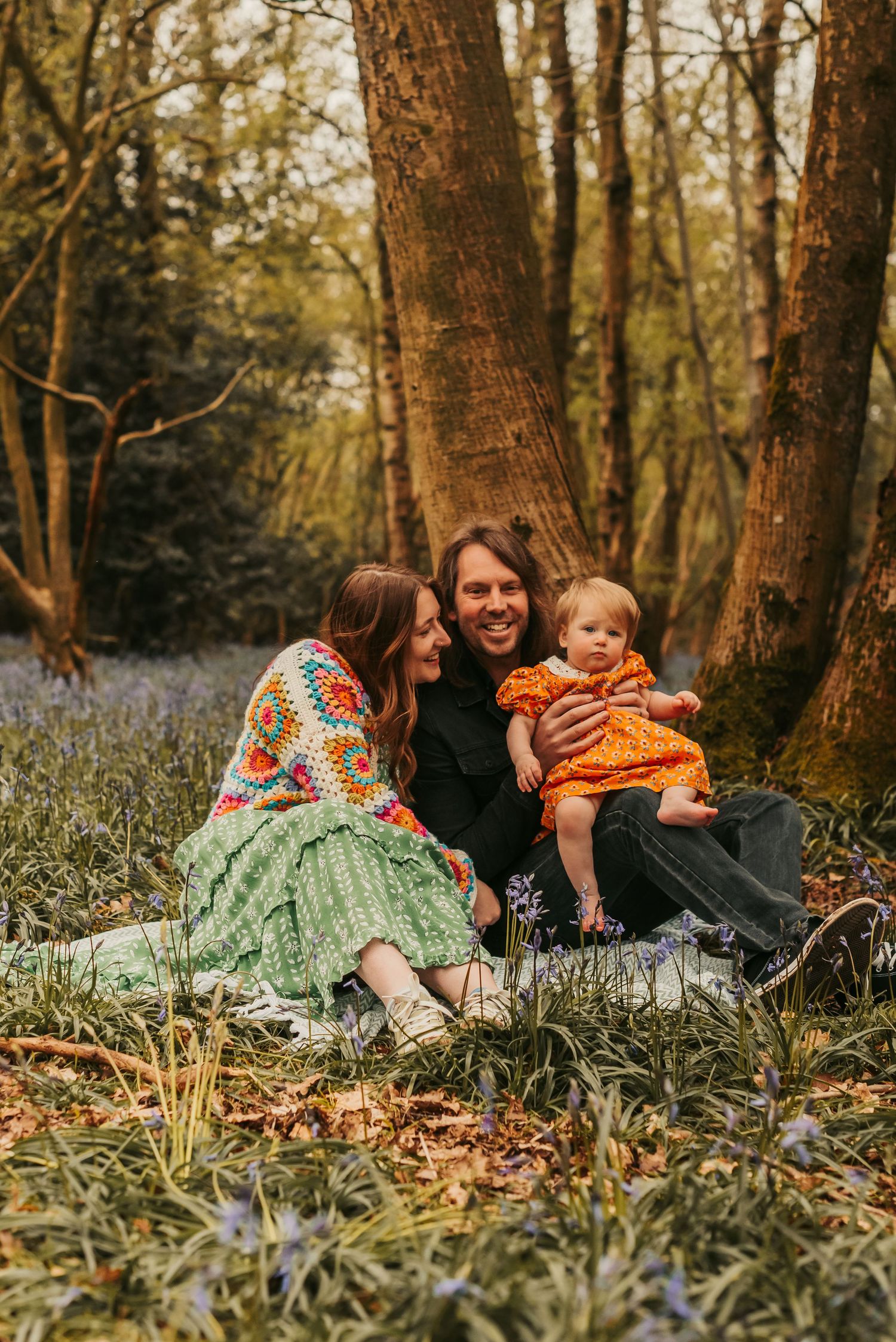 A family sits together on the forest floor surrounded by bluebells during golden hour in a wooded setting.