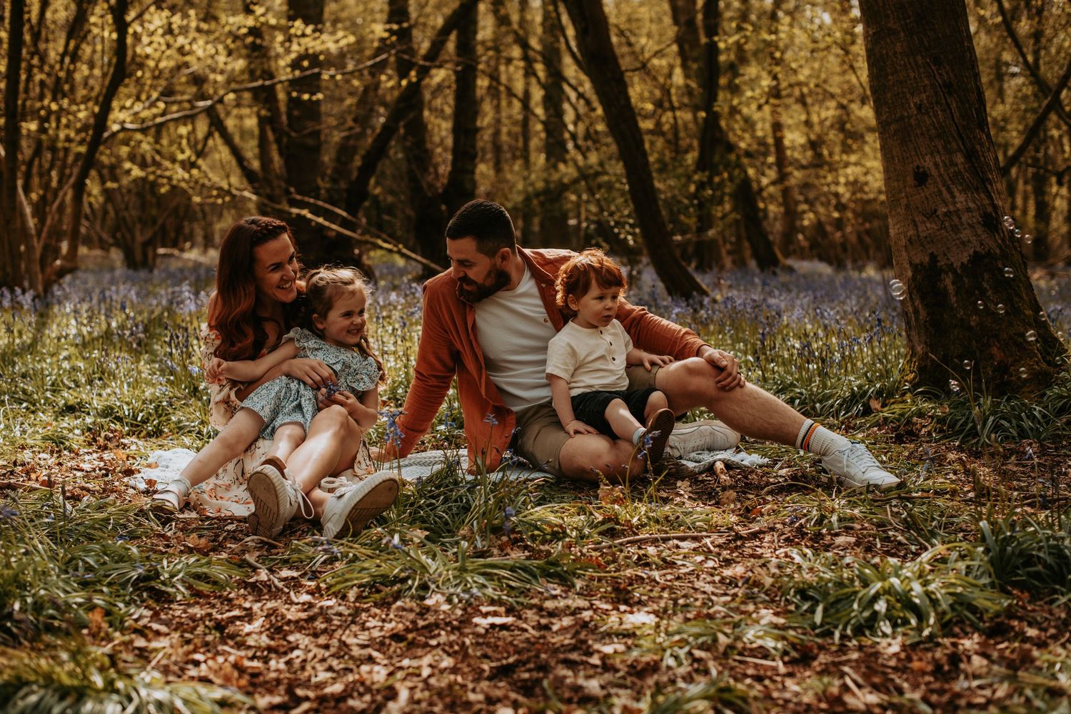 A family relaxes together on autumn leaves in a forest with blooming bluebells during golden hour.