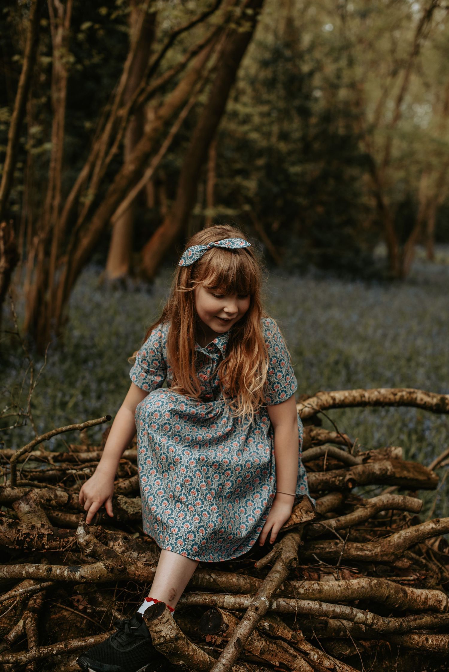 A girl in a patterned dress explores fallen logs in a mystical bluebell forest.