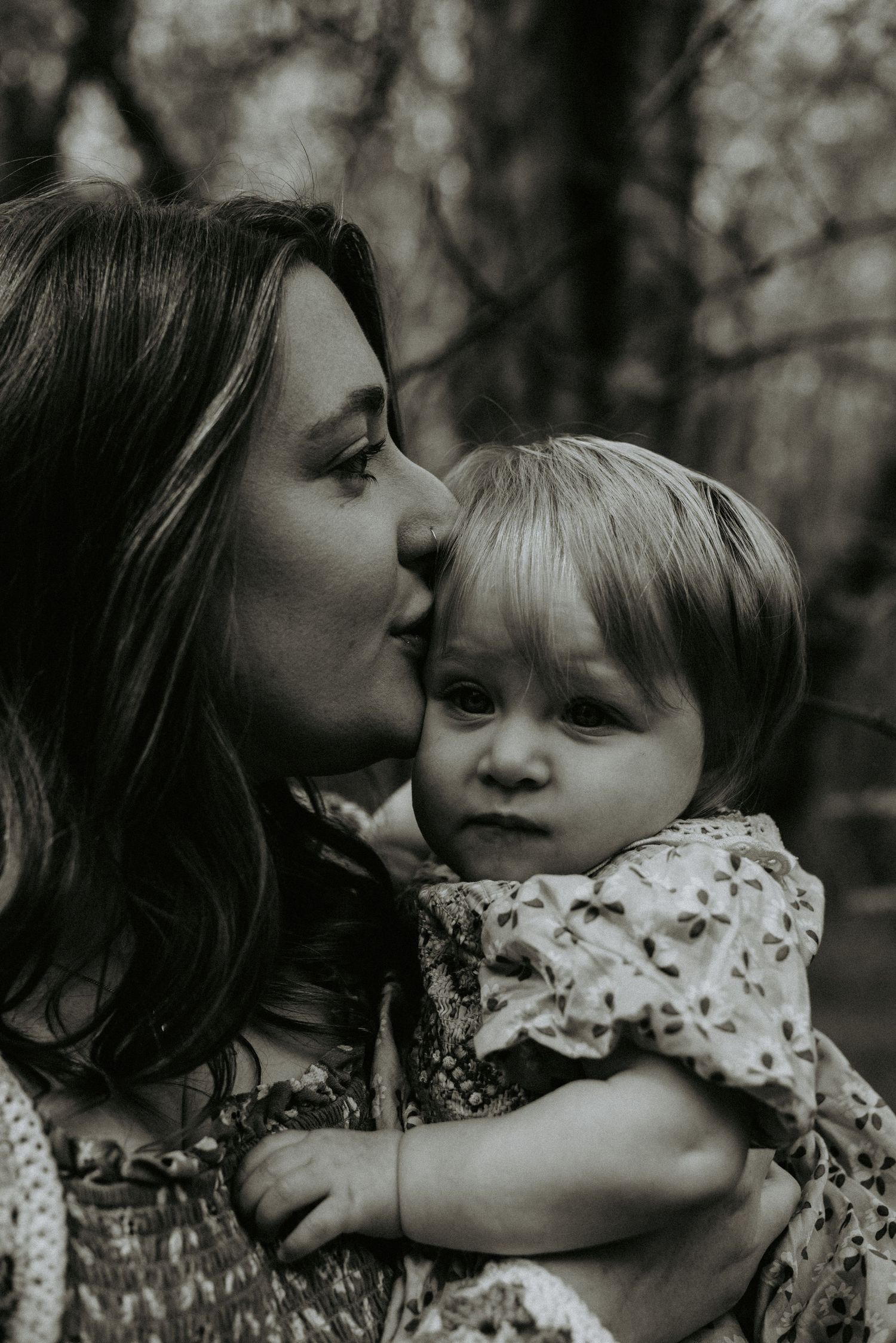 A tender black and white portrait of a mother kissing her young toddler's head in a forest setting.