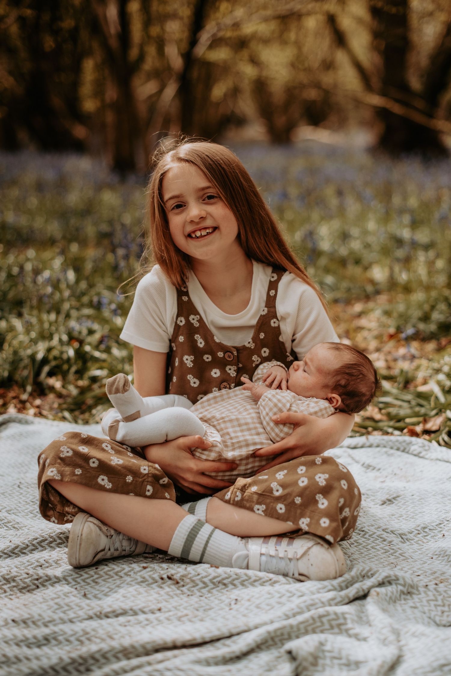 In a forest of bluebells, someone wearing brown polka dot overalls sits on a blanket holding a baby on their lap.
